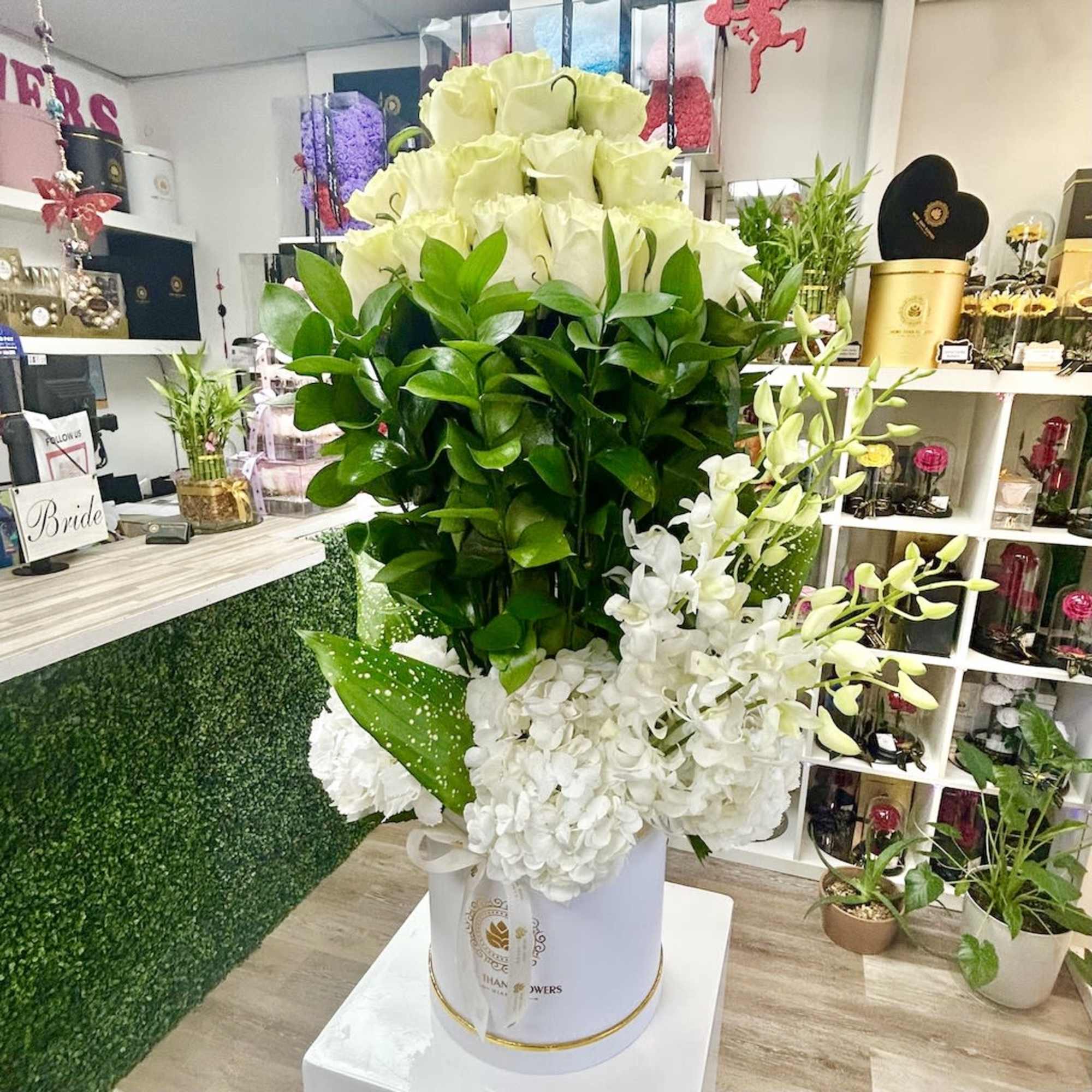 Tall white hatbox arrangement with white roses, hydrangeas, and orchids above dense green foliage