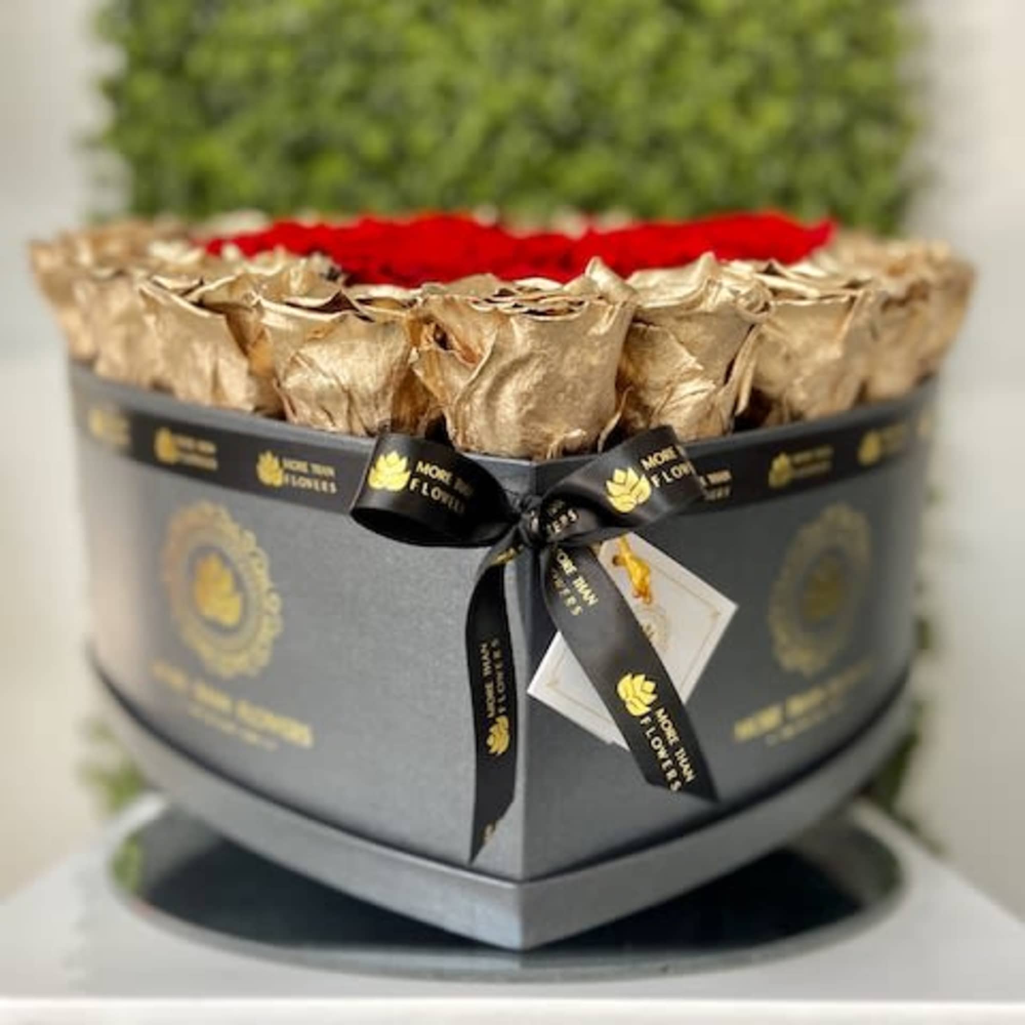 Heart-shaped black box filled with red roses framed by gold-wrapped blooms and tied with a black ribbon.