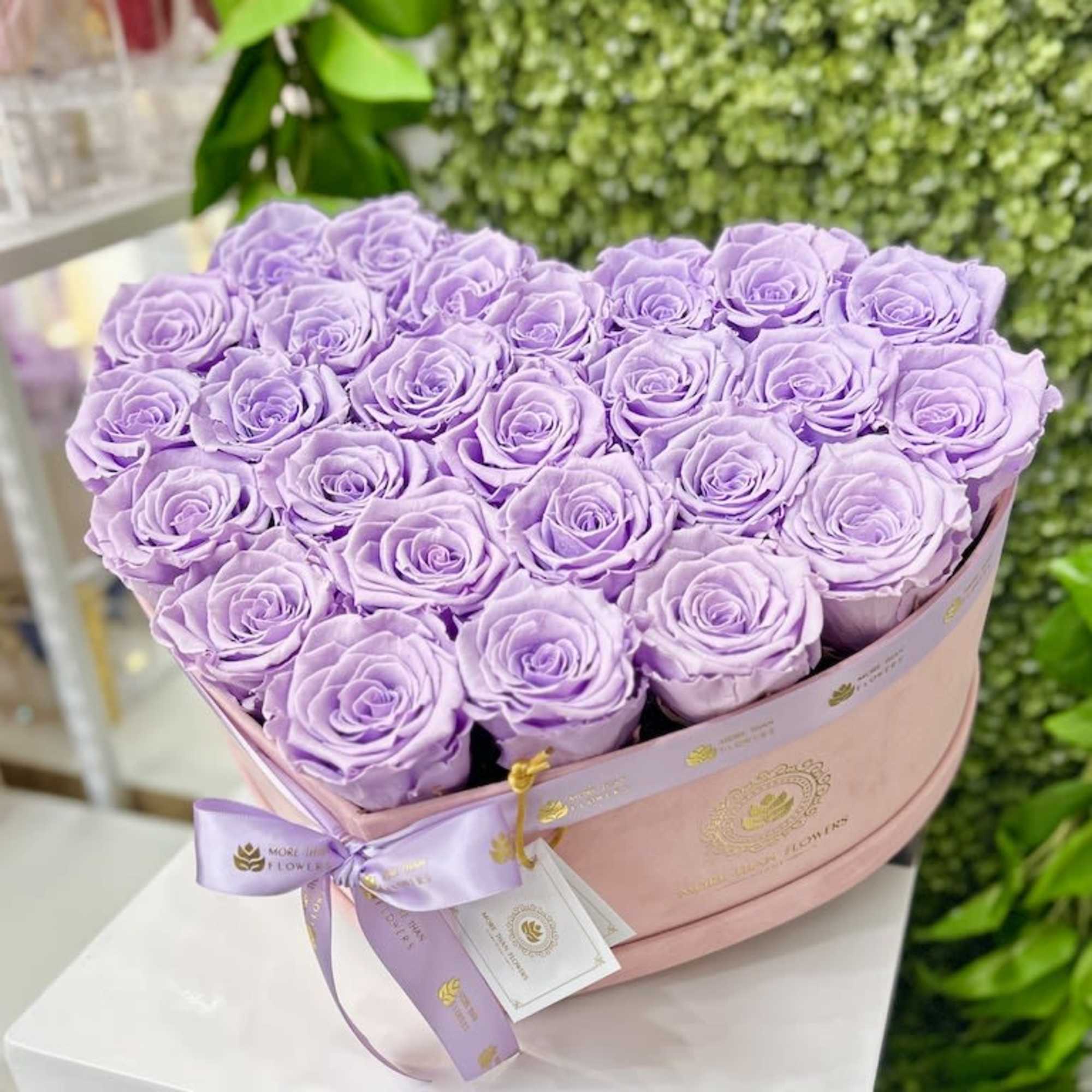 Heart-shaped box of tightly packed lavender roses with a pink band and ribbon on a white pedestal.