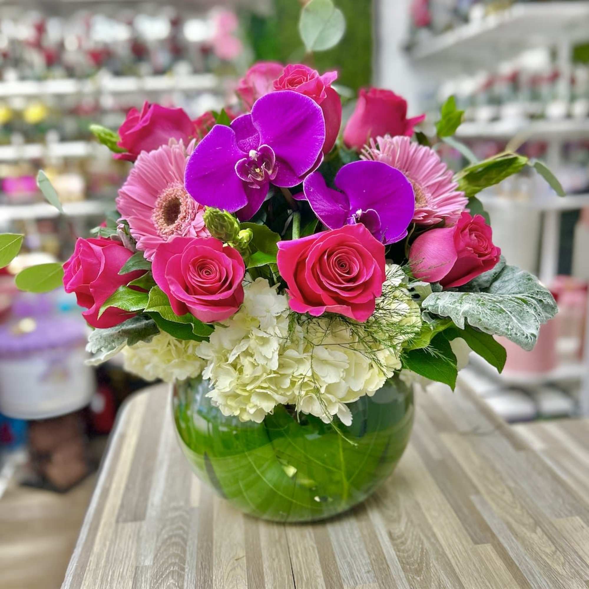 Low round arrangement of hot pink roses, purple orchids and white hydrangea in a green-lined glass bowl