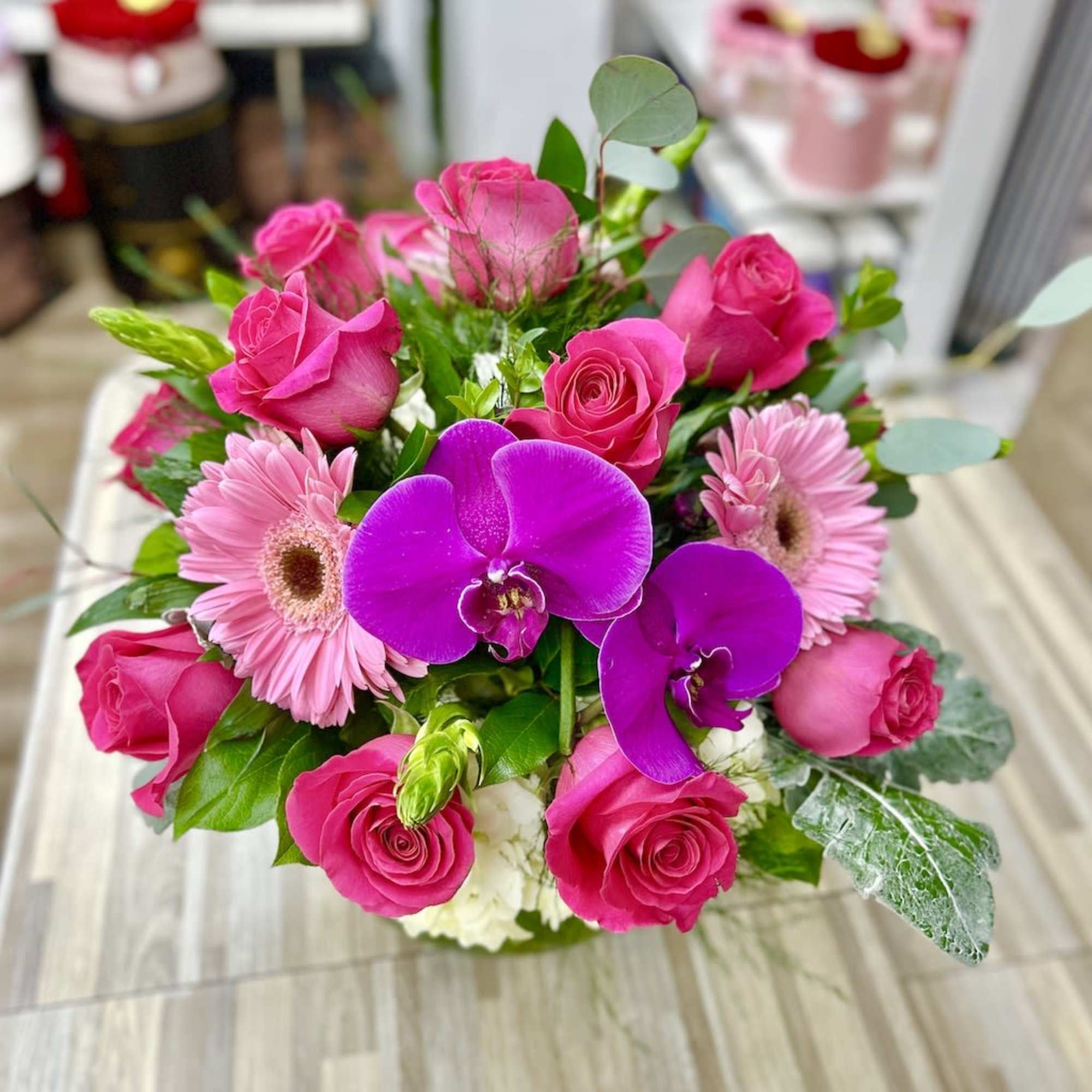 Low arrangement of hot pink roses, pink gerbera daisies, and purple orchids in a compact vase