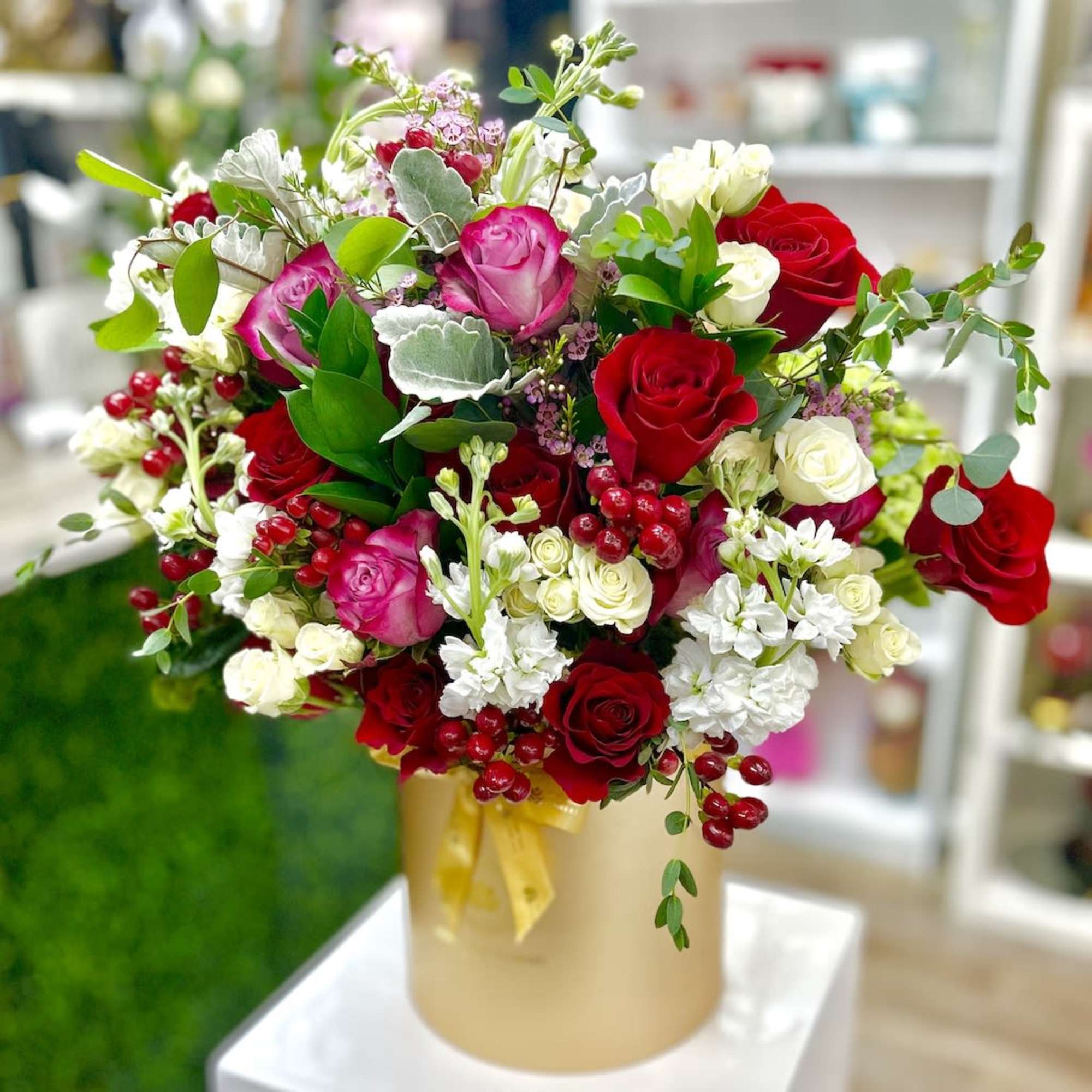 Round arrangement of red, pink, and white roses with berries in a gold hatbox-style container