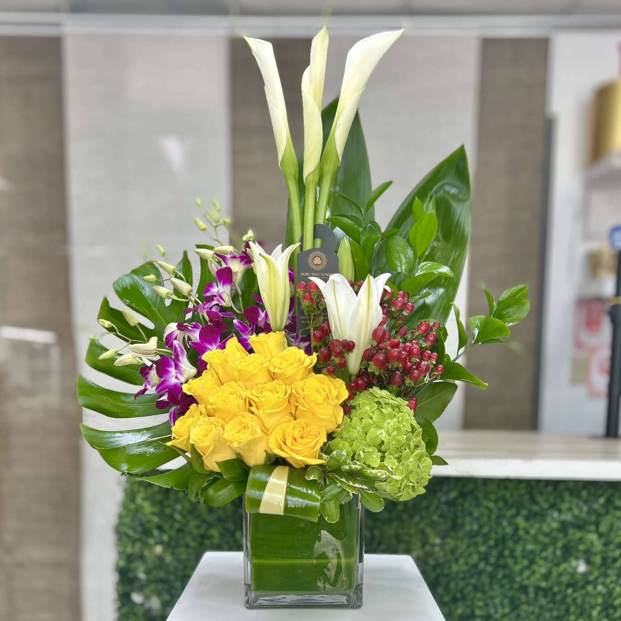 Tall arrangement with yellow roses, white lilies, orchids and calla lilies in a glass vase with large tropical leaves