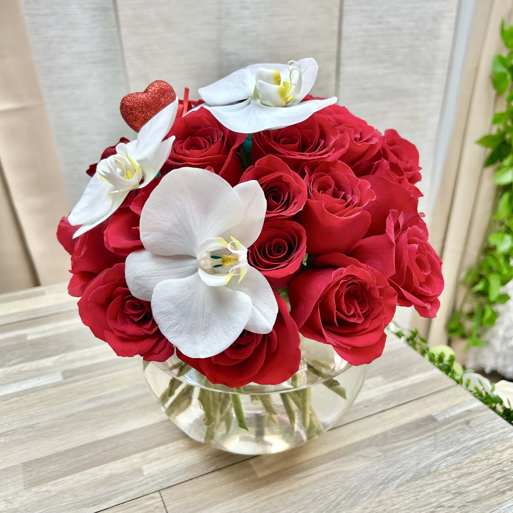 Round glass vase of red roses with white orchids and a small red heart pick.