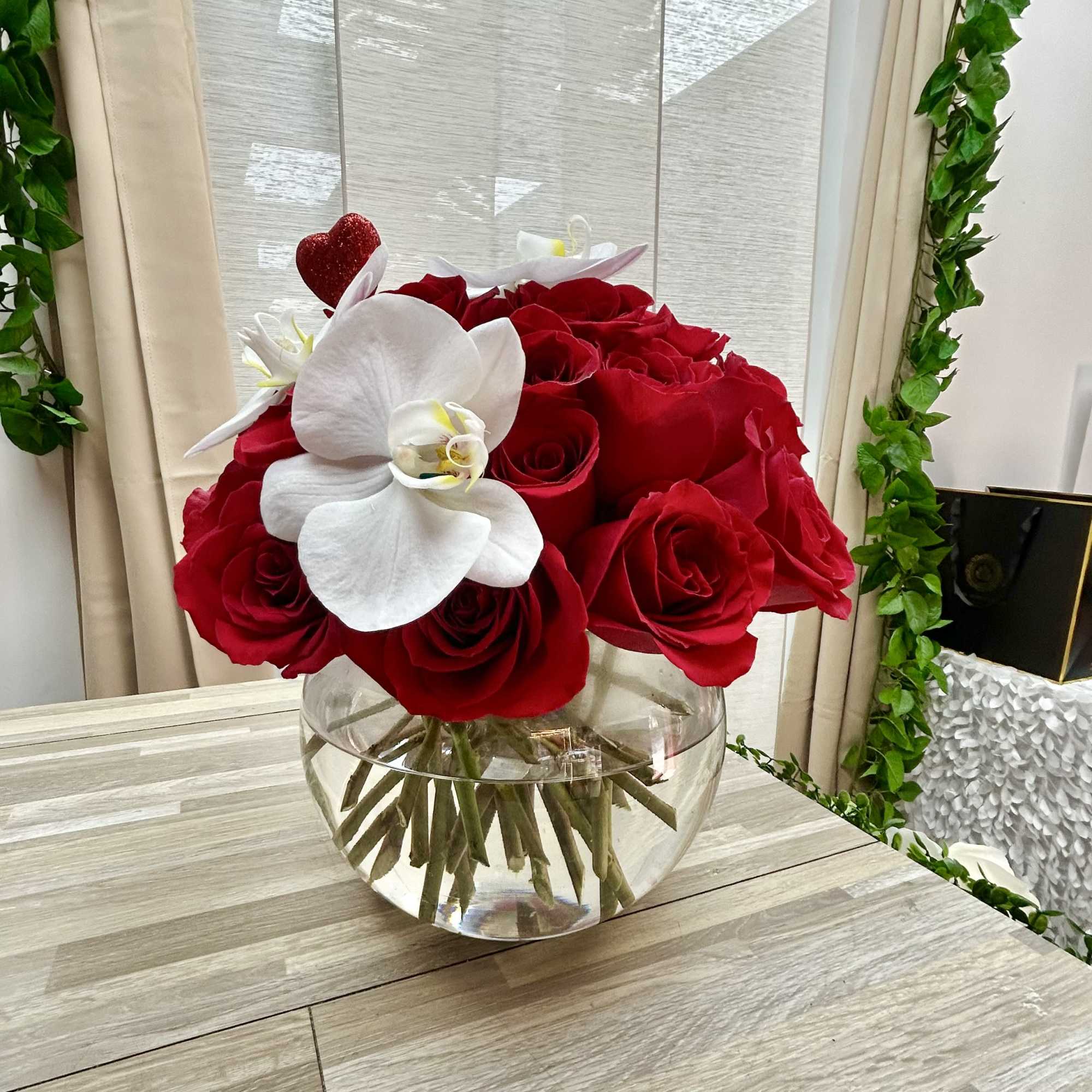 Low round arrangement of red roses with white orchids in a clear glass bowl vase and a red heart pick