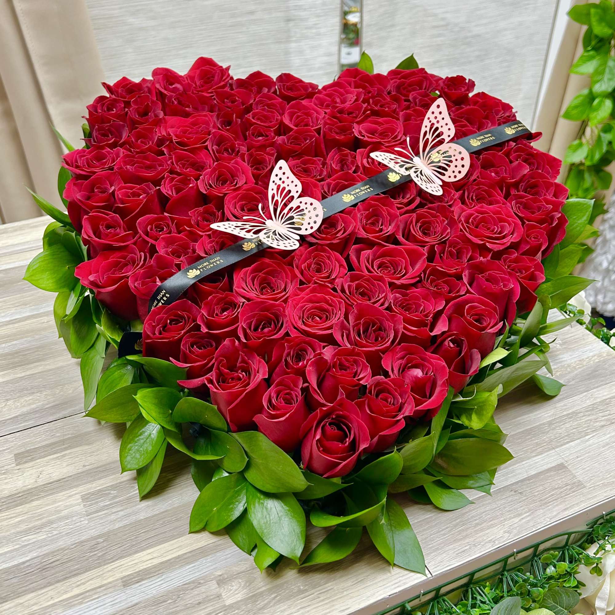 Heart-shaped arrangement of red roses edged with green foliage and butterfly accents