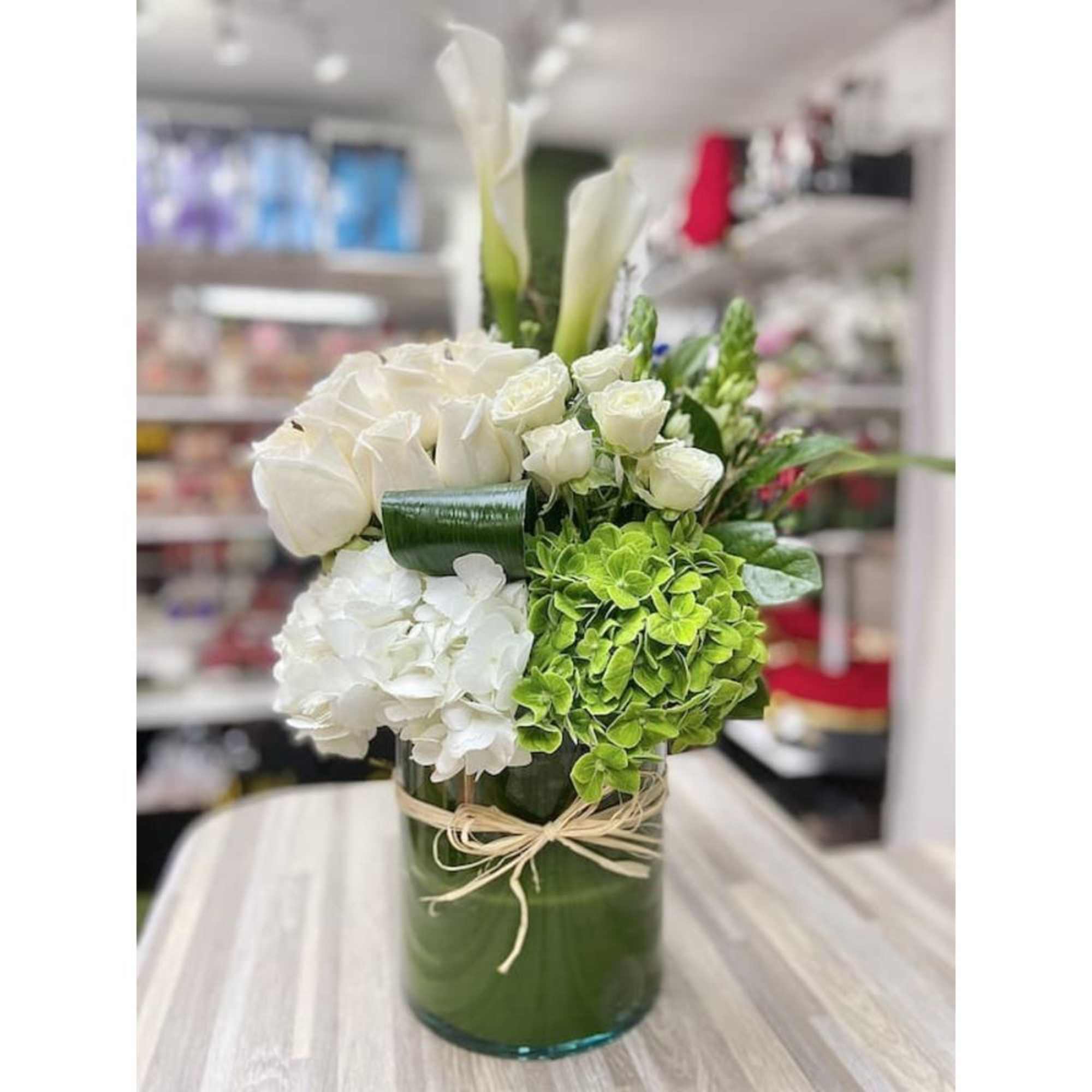White roses, calla lilies, and green hydrangeas arranged in a glass cylinder vase tied with raffia