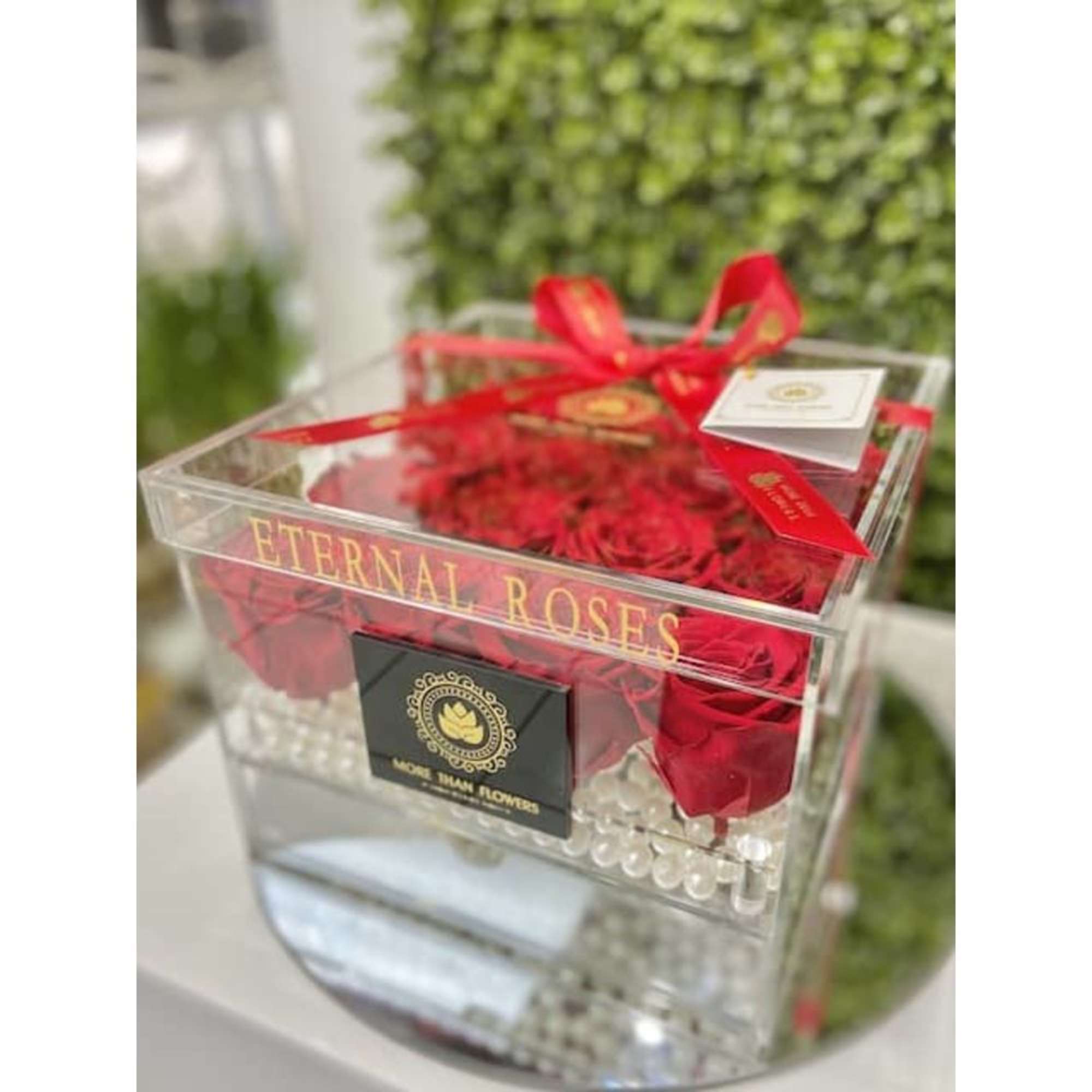 Clear acrylic box filled with red roses and pearl accents, tied with a red ribbon and card.