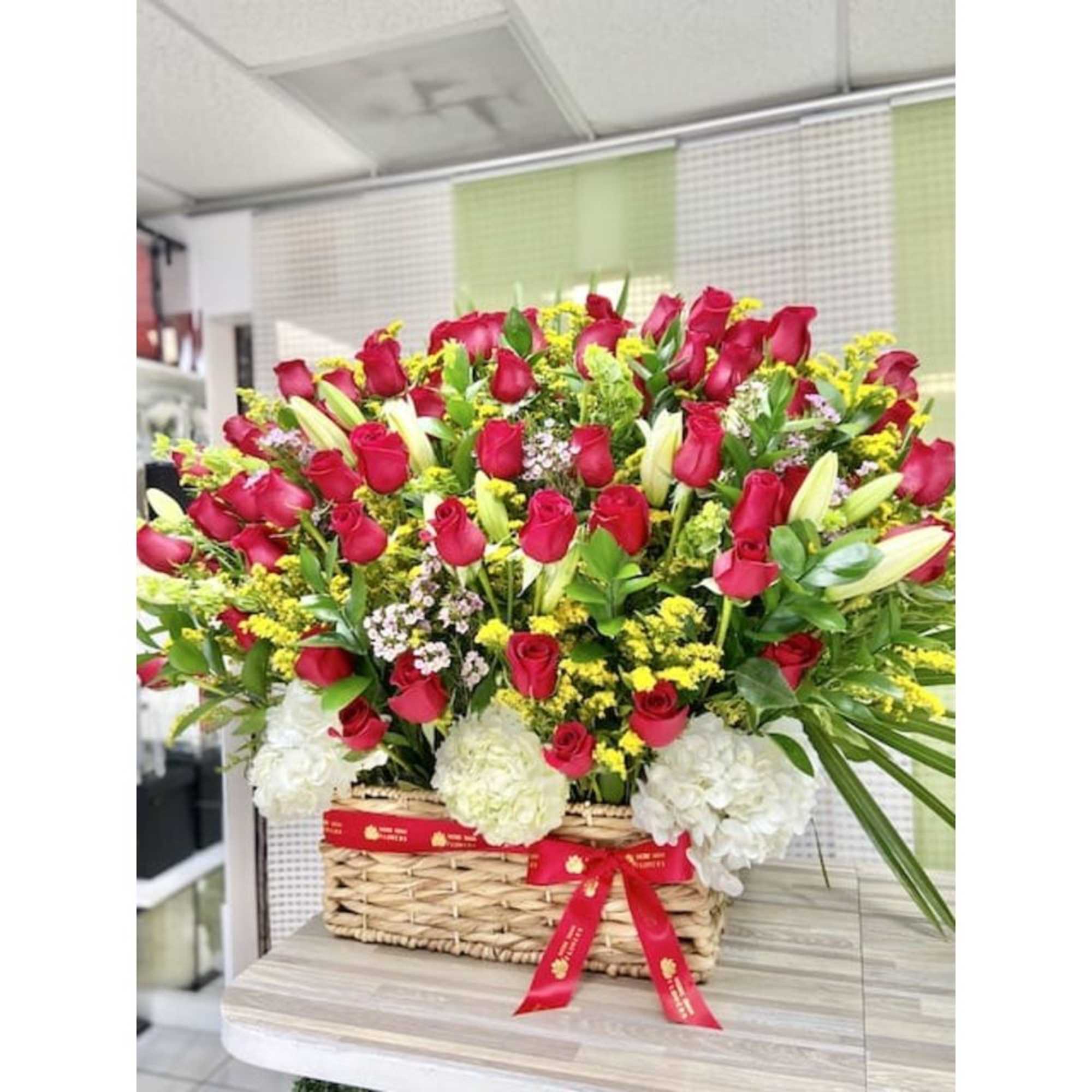 Large basket arrangement of red roses, white lilies, yellow accents, and white hydrangeas with a red ribbon