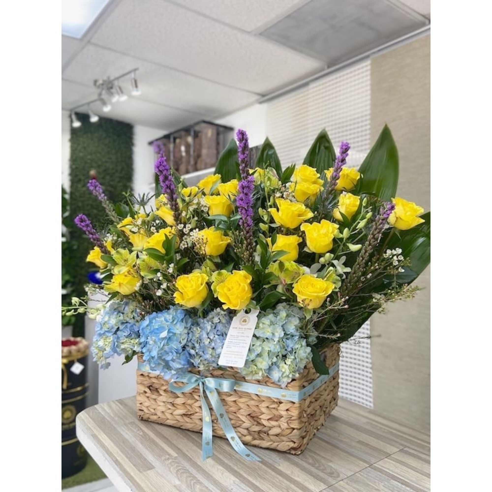 Basket arrangement with yellow roses, purple spikes, and blue hydrangeas tied with a blue ribbon