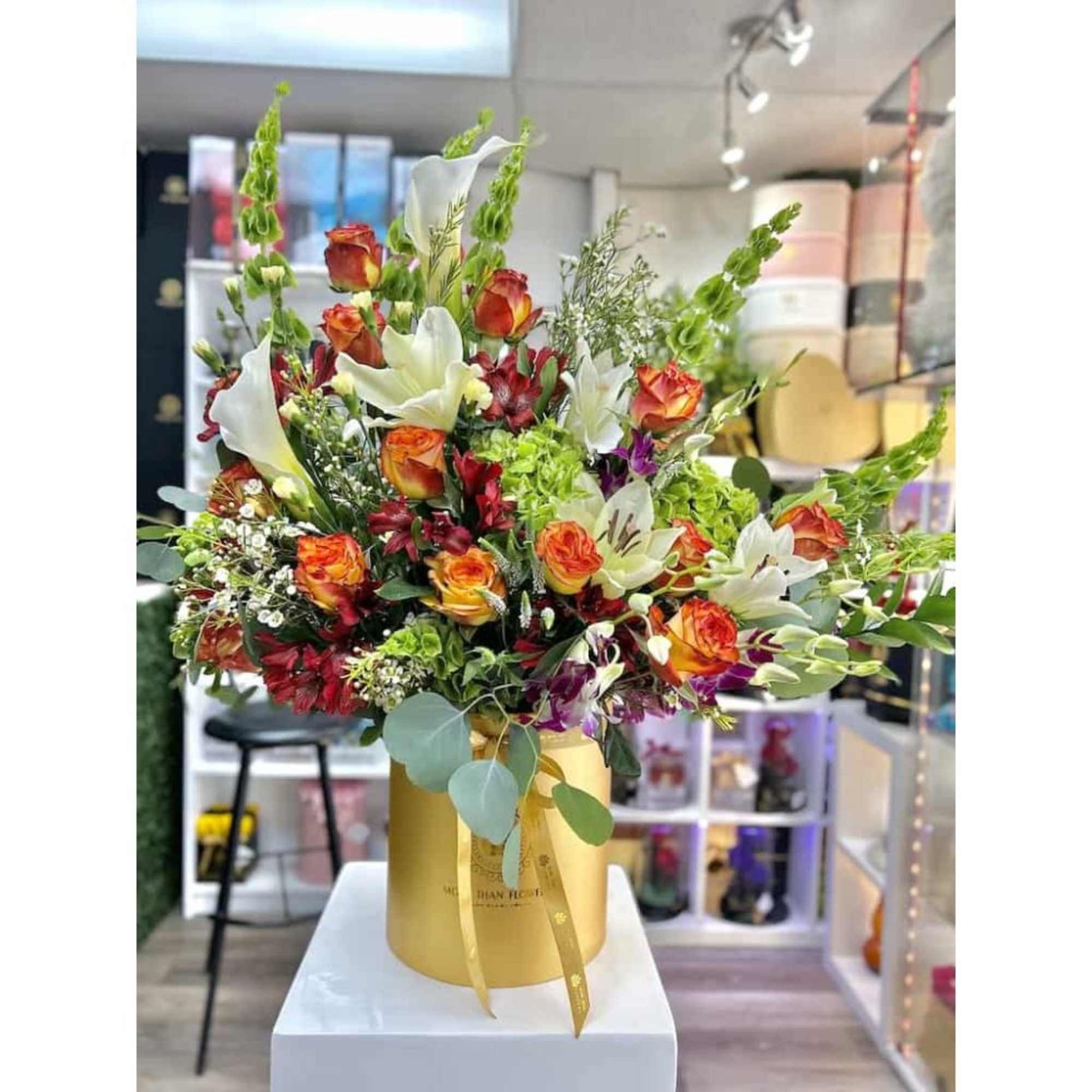 Large mixed arrangement of orange roses, white lilies, and assorted blooms in a gold hatbox container.