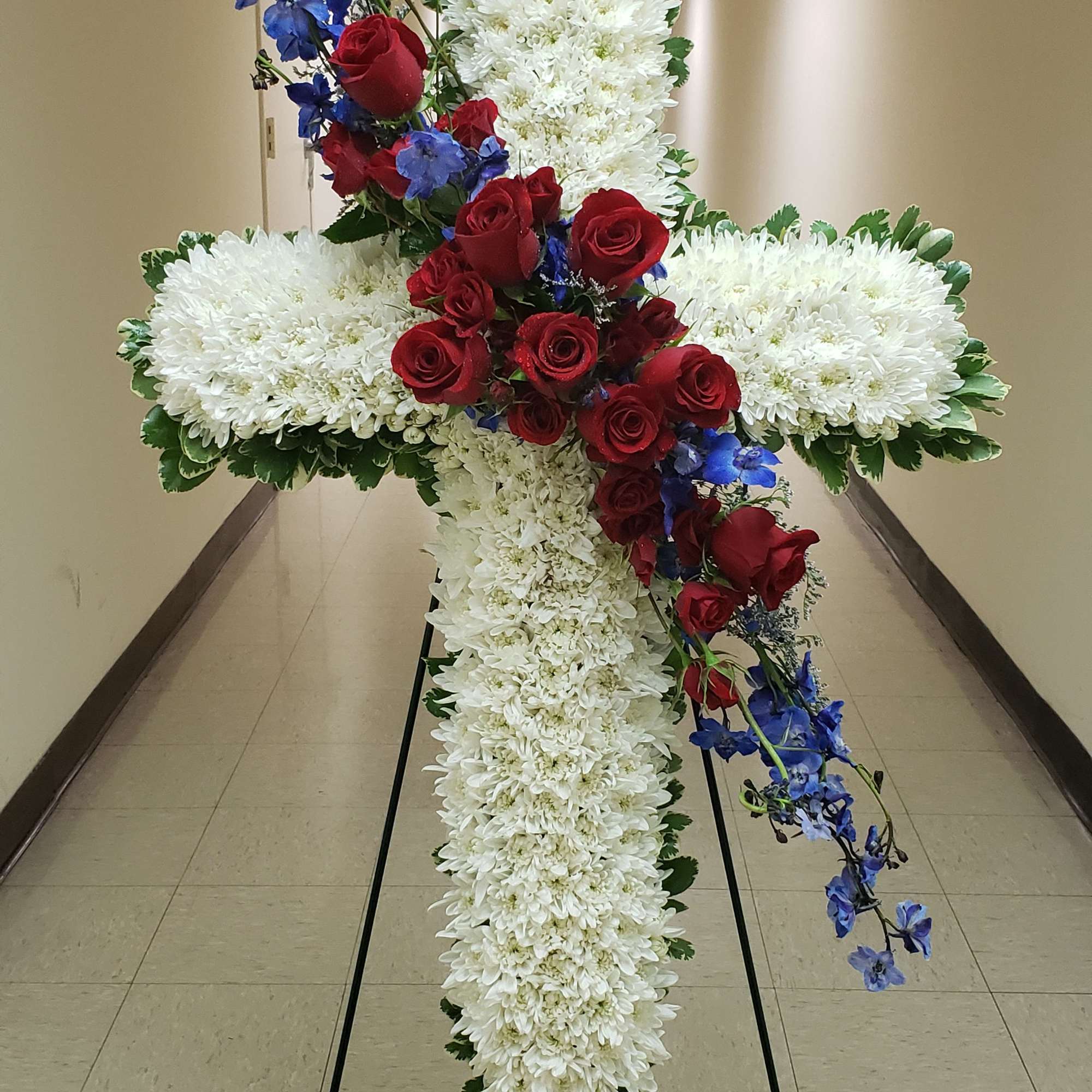 Standing cross-shaped spray of white flowers with red roses and blue blooms on an easel