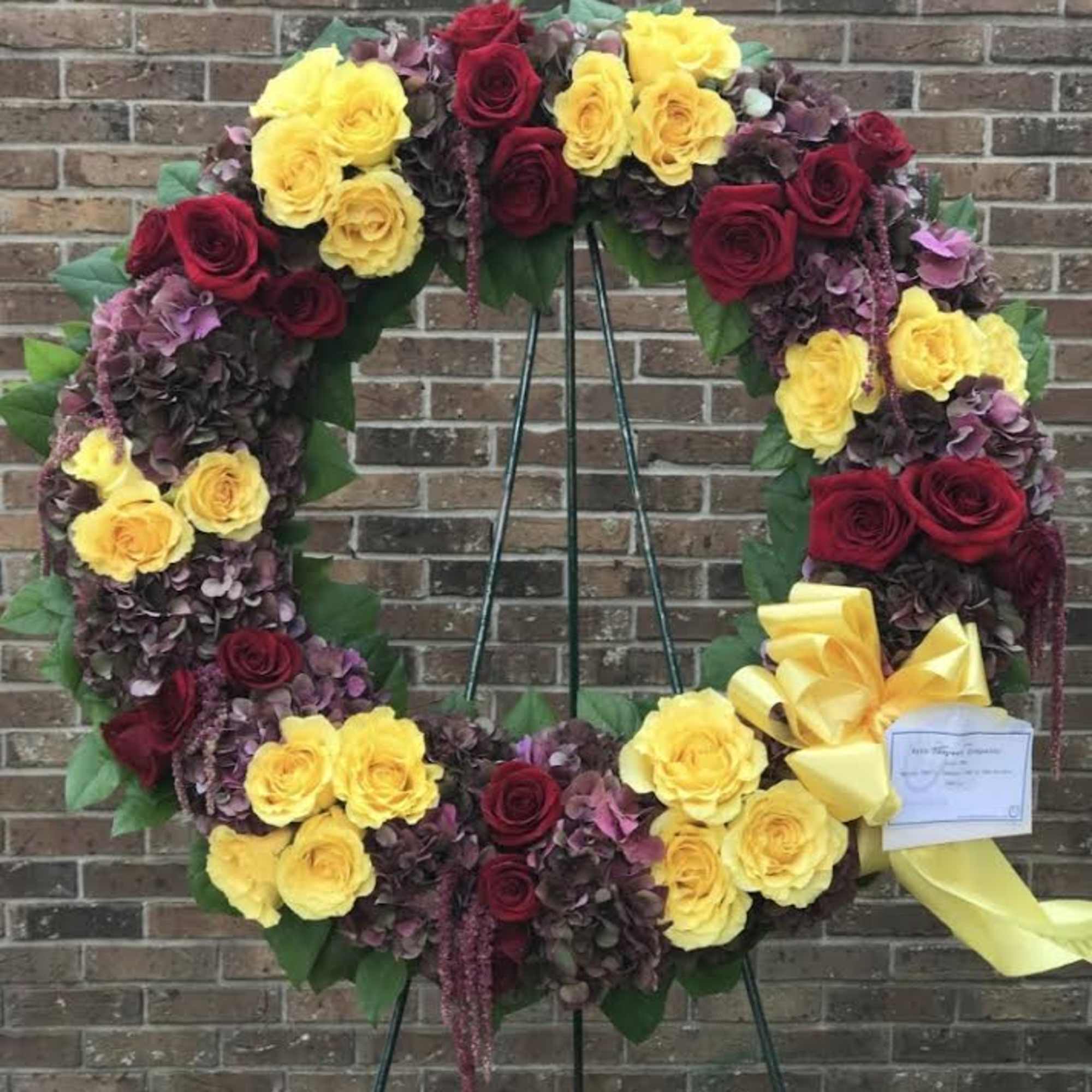 Standing wreath of yellow and red roses with burgundy hydrangeas and yellow bow on metal easel