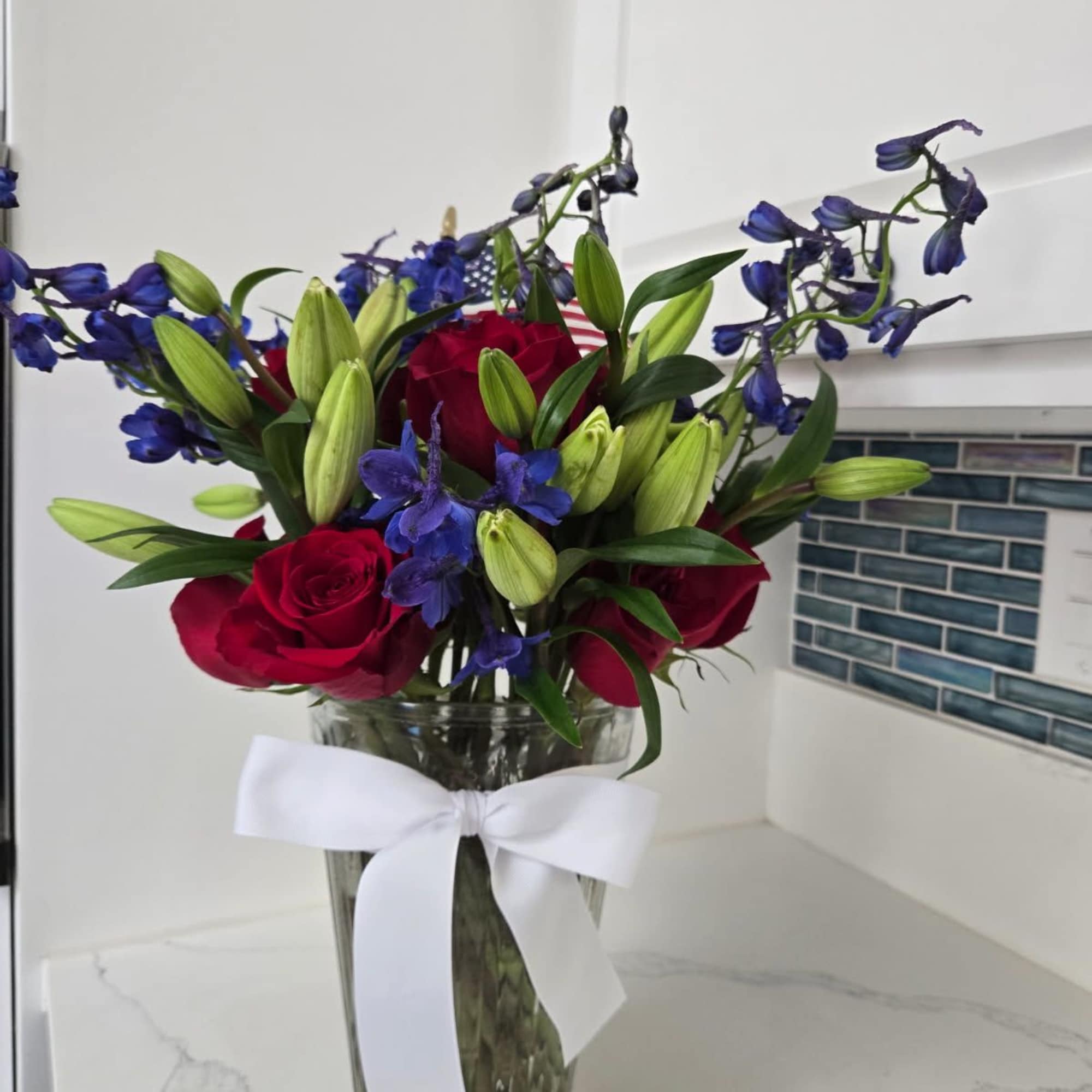 Red roses and blue flowers in a clear glass vase with a white ribbon