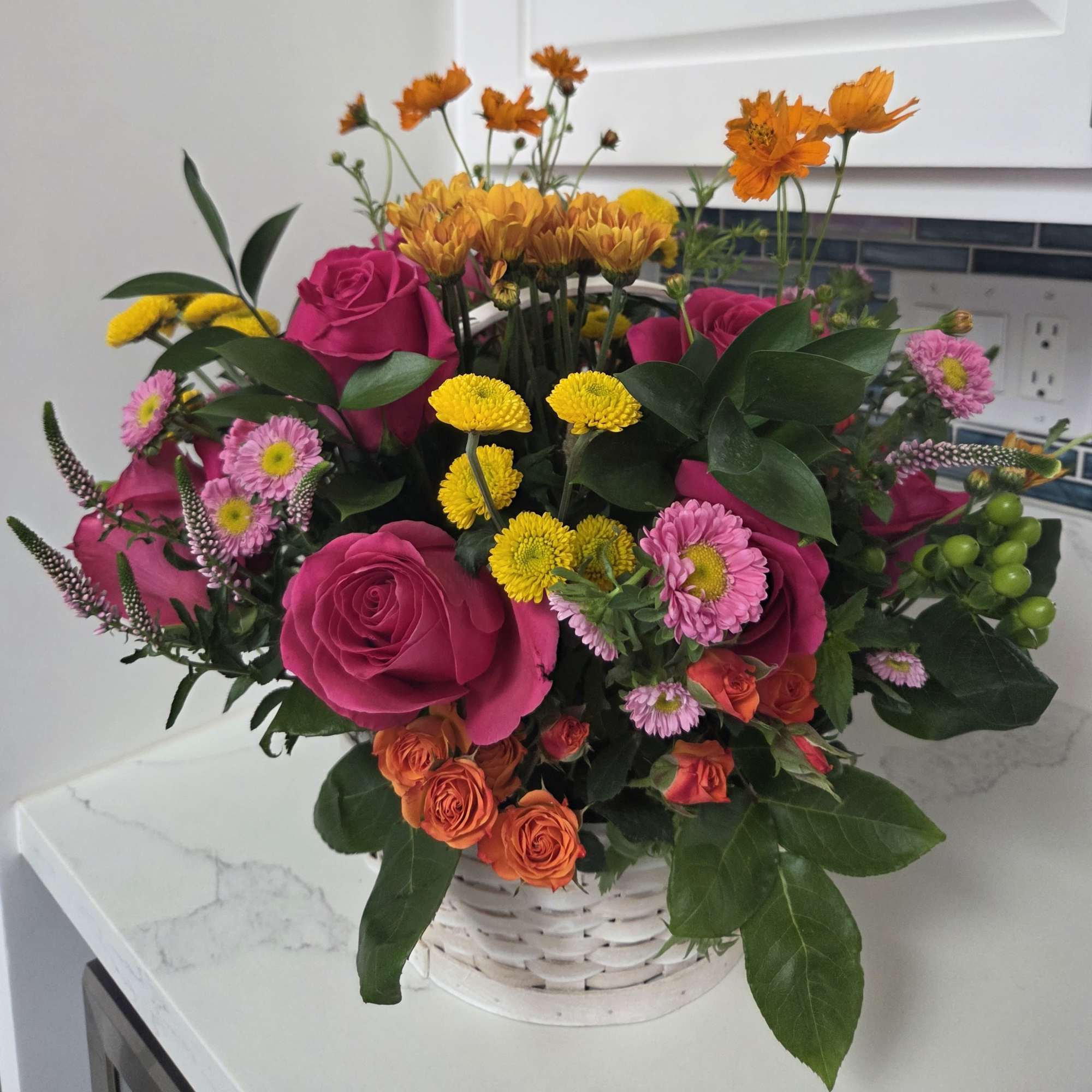 Mixed bouquet of pink roses, yellow mums, and orange flowers in a white basket
