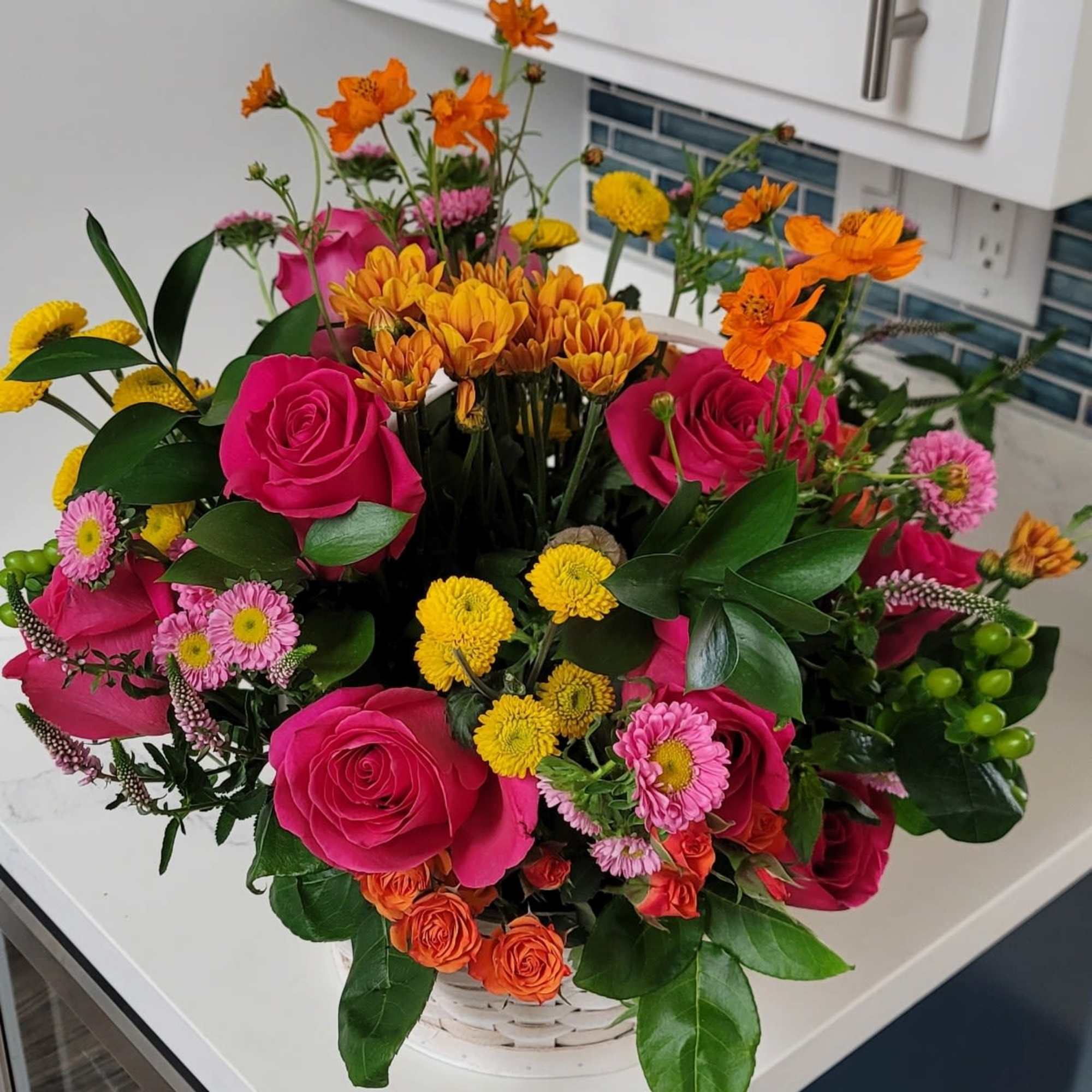 Mixed bouquet of pink roses, orange blooms, and yellow flowers in a white basket