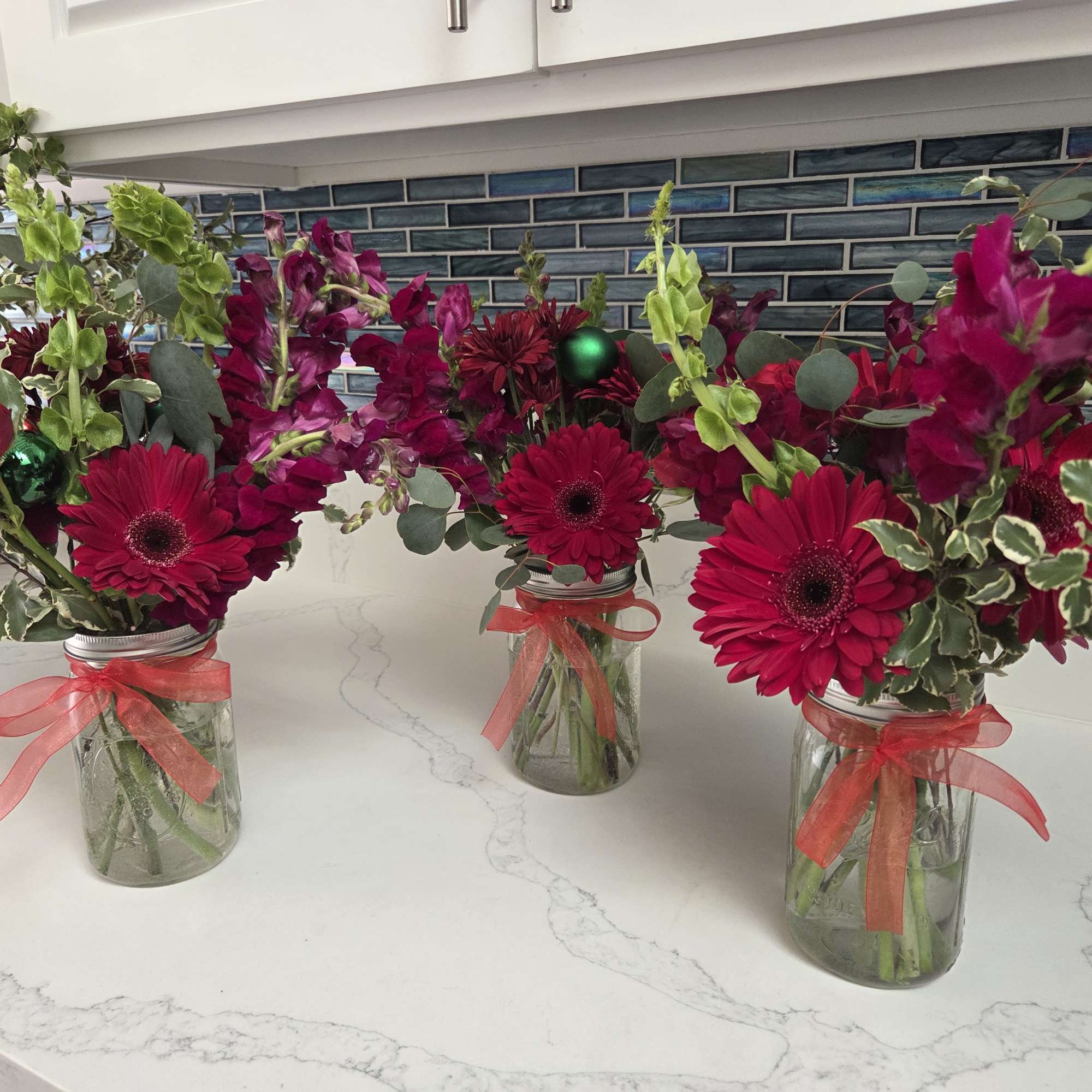 Three small floral arrangements in glass jars with bright pink blooms and ribbons.
