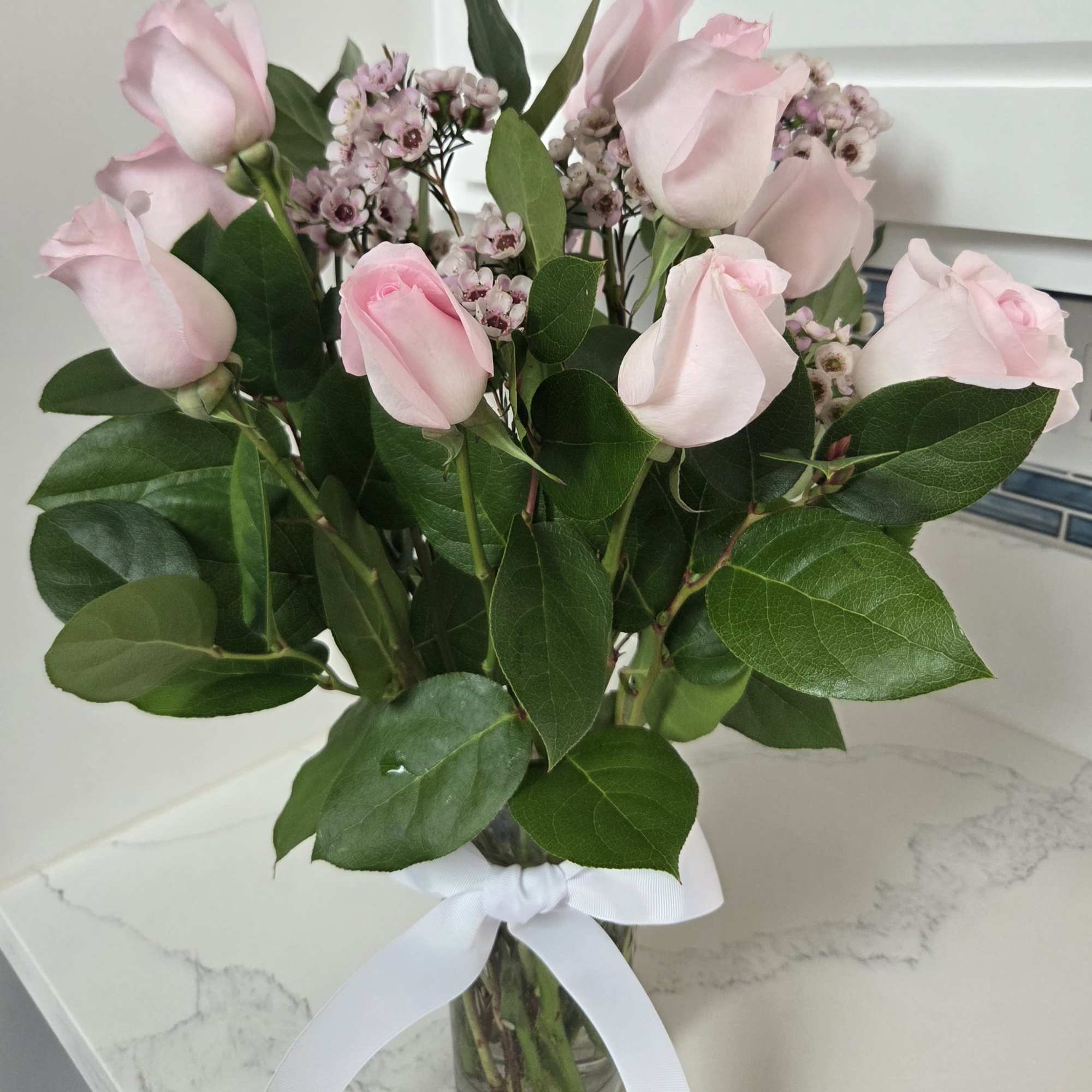 Pink roses and small waxflower blooms in a glass vase with a white ribbon.