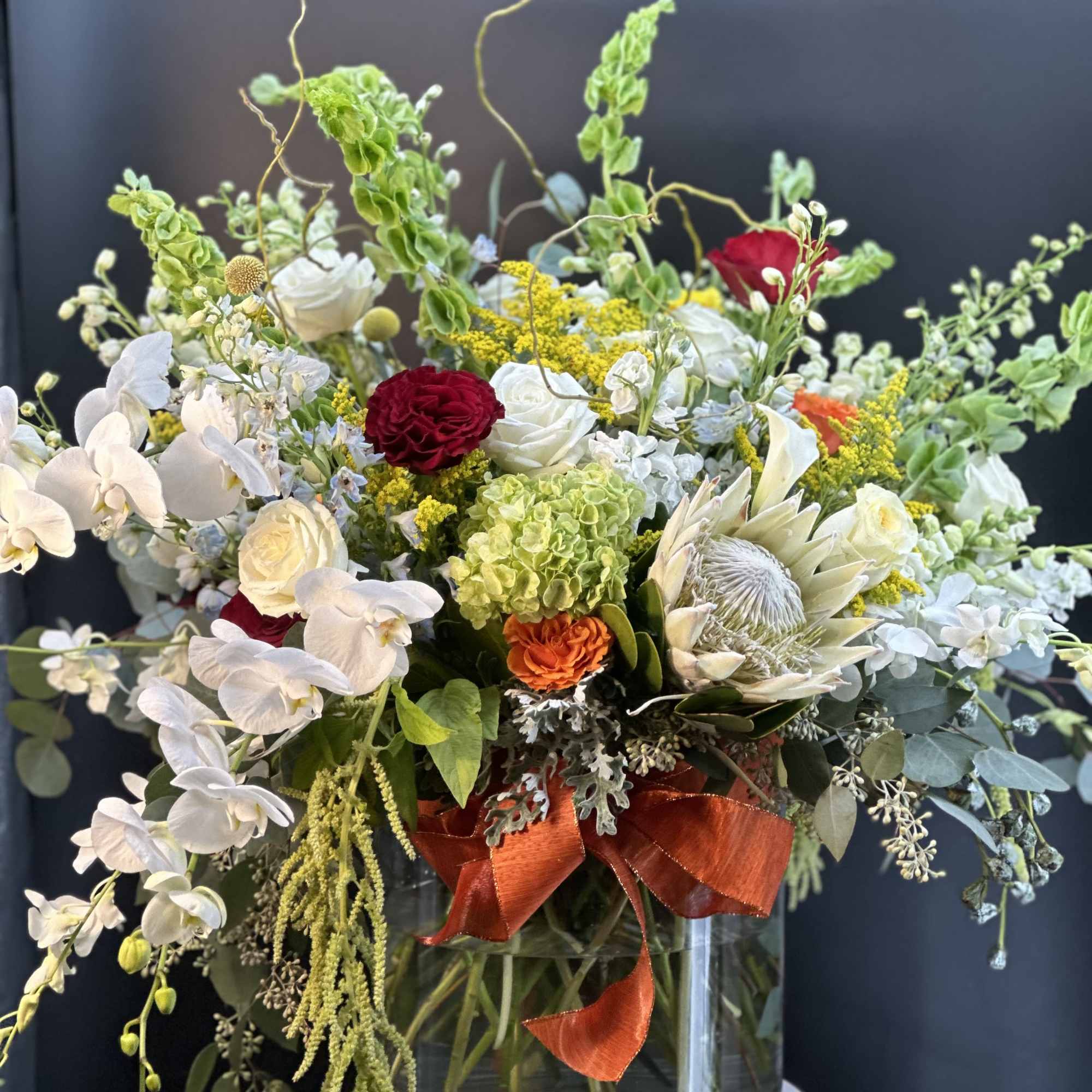 Large mixed arrangement with white orchids, roses, green hydrangea, red and orange blooms in a clear glass vase.