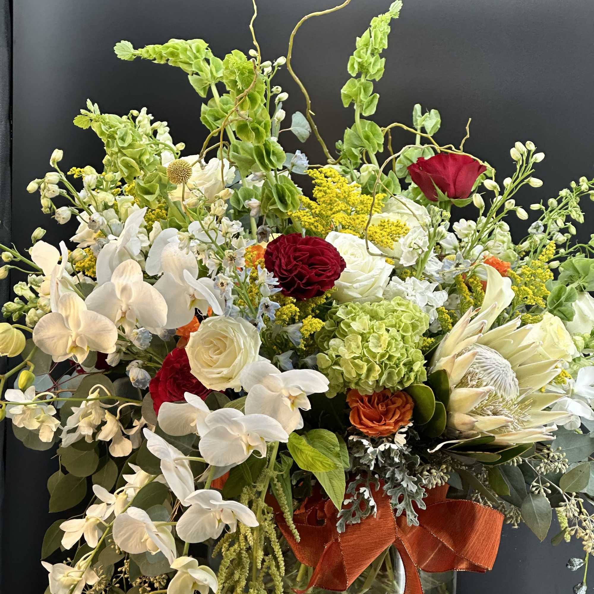 Tall mixed arrangement with white orchids, roses, green hydrangea and protea in a clear glass vase.