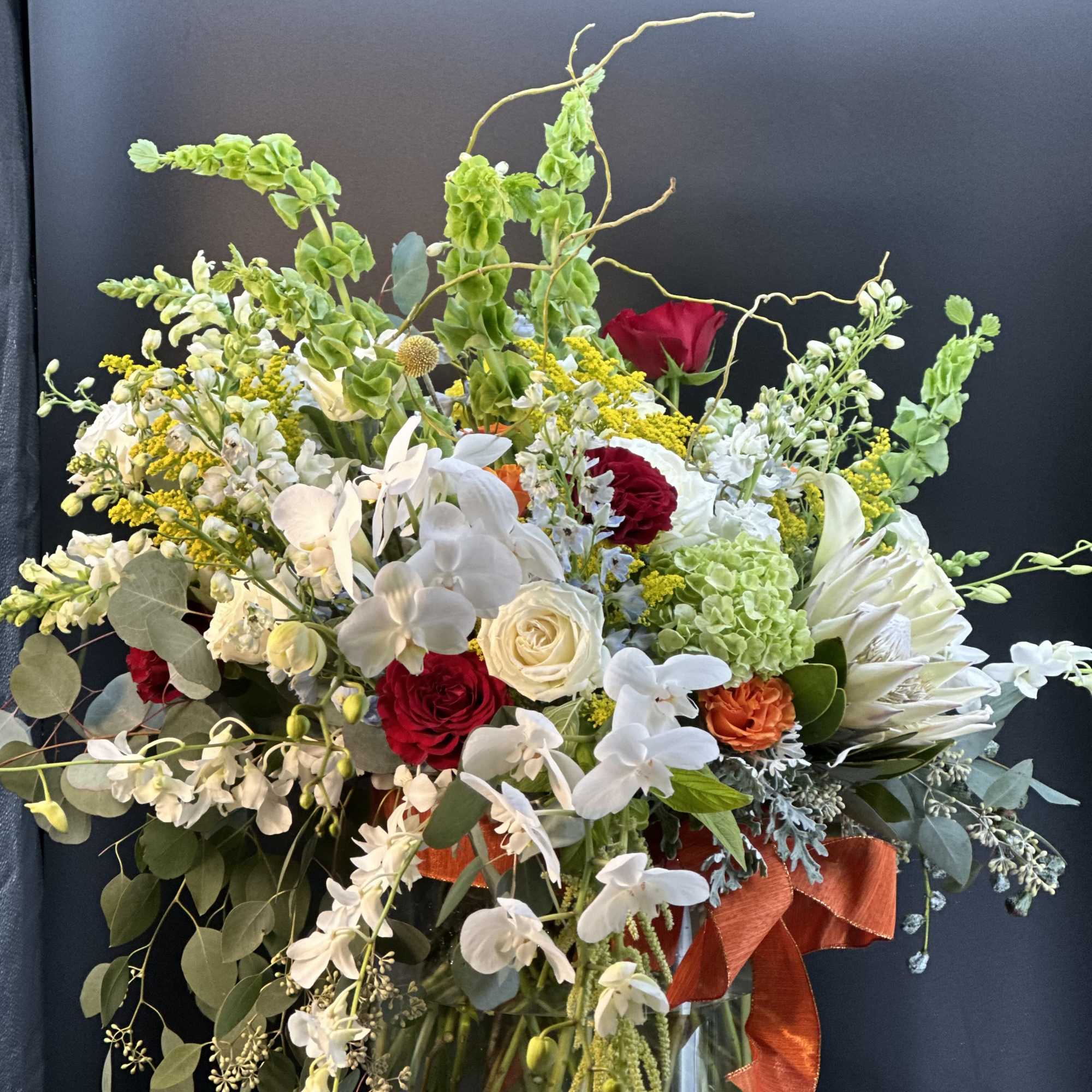 Large mixed arrangement of white orchids, red and white roses, and protea in a glass vase with orange ribbon