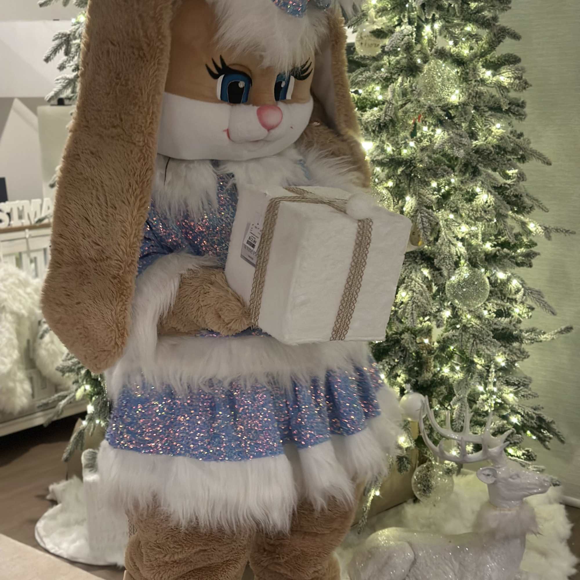 Large plush bunny in a sparkly blue outfit holding a wrapped gift beside lit Christmas trees.