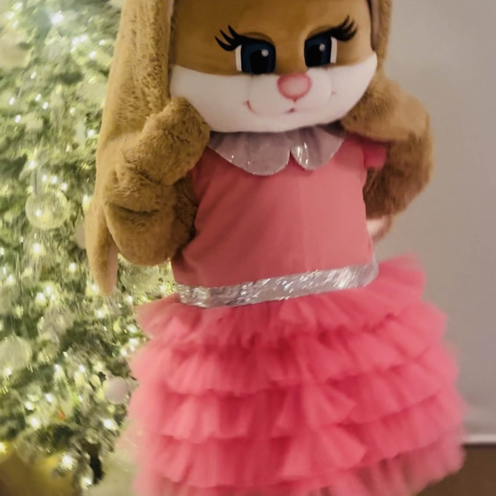 Large plush bunny in a pink ruffled dress standing beside a lit Christmas tree.