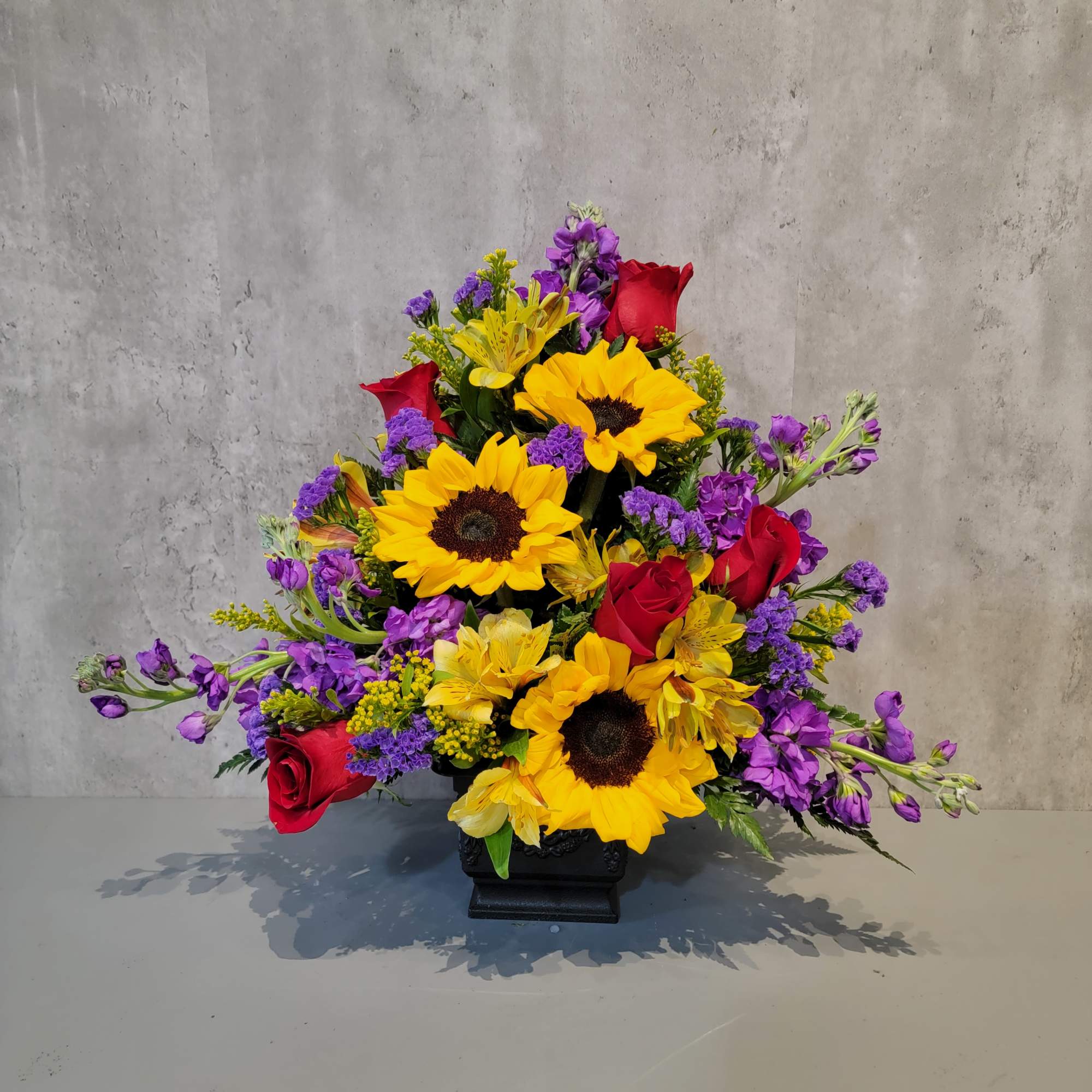 Colorful floral arrangement with sunflowers, red roses, and purple blooms in a black vase