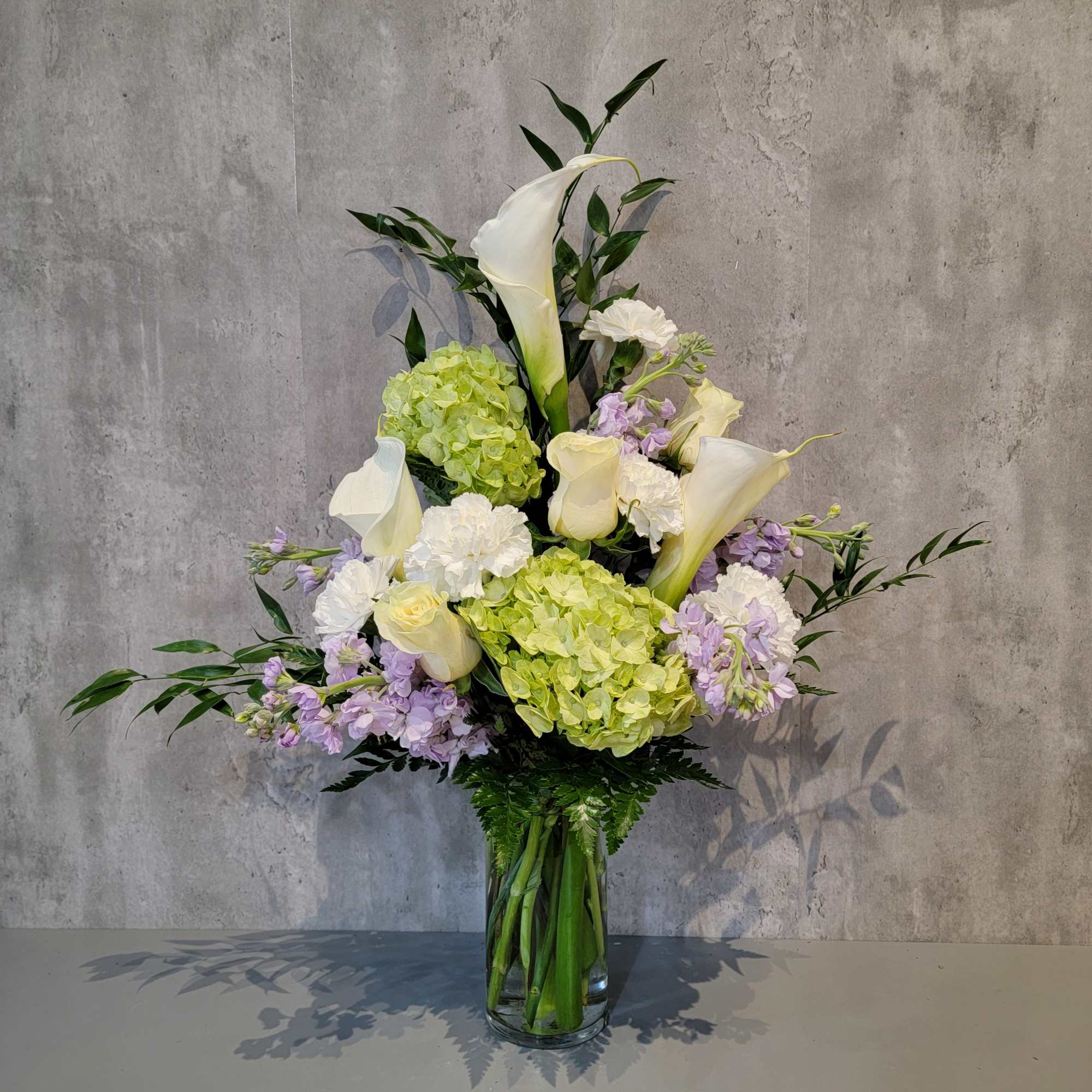 Tall bouquet of white calla lilies, hydrangeas, and lavender flowers in a glass vase