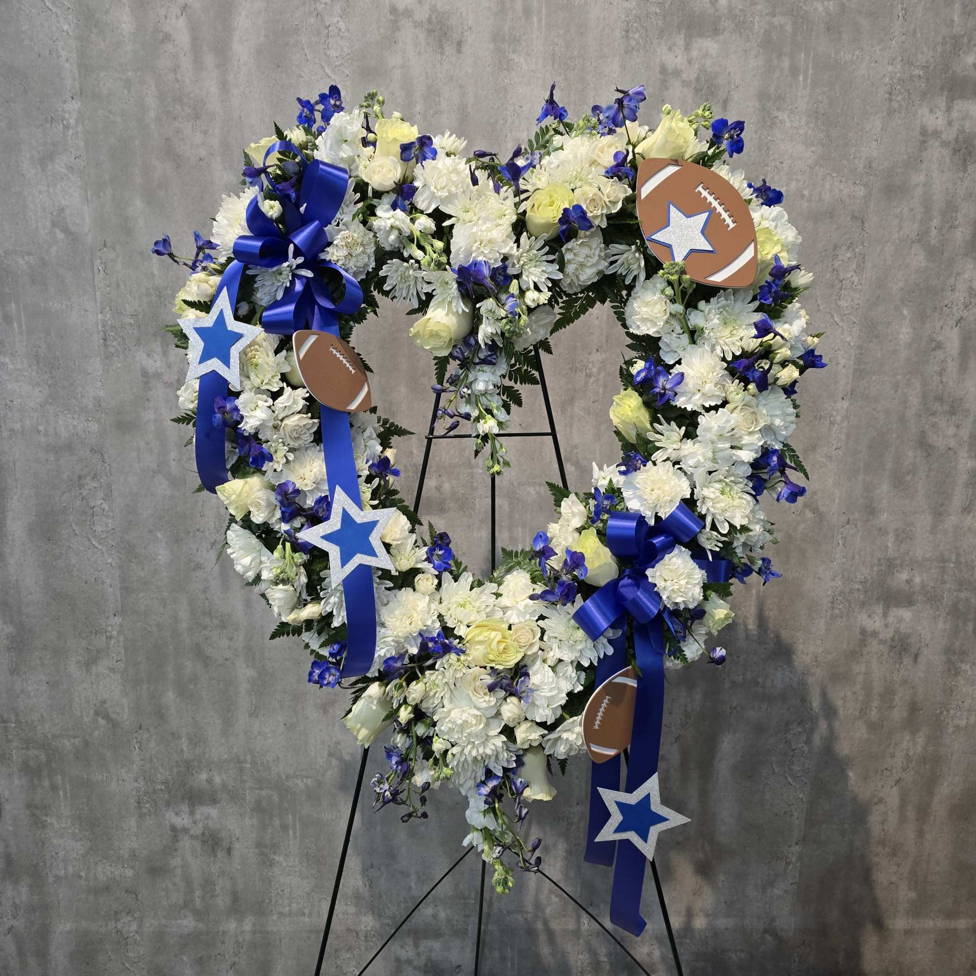 Heart-shaped floral wreath with blue ribbons and football decorations on a stand