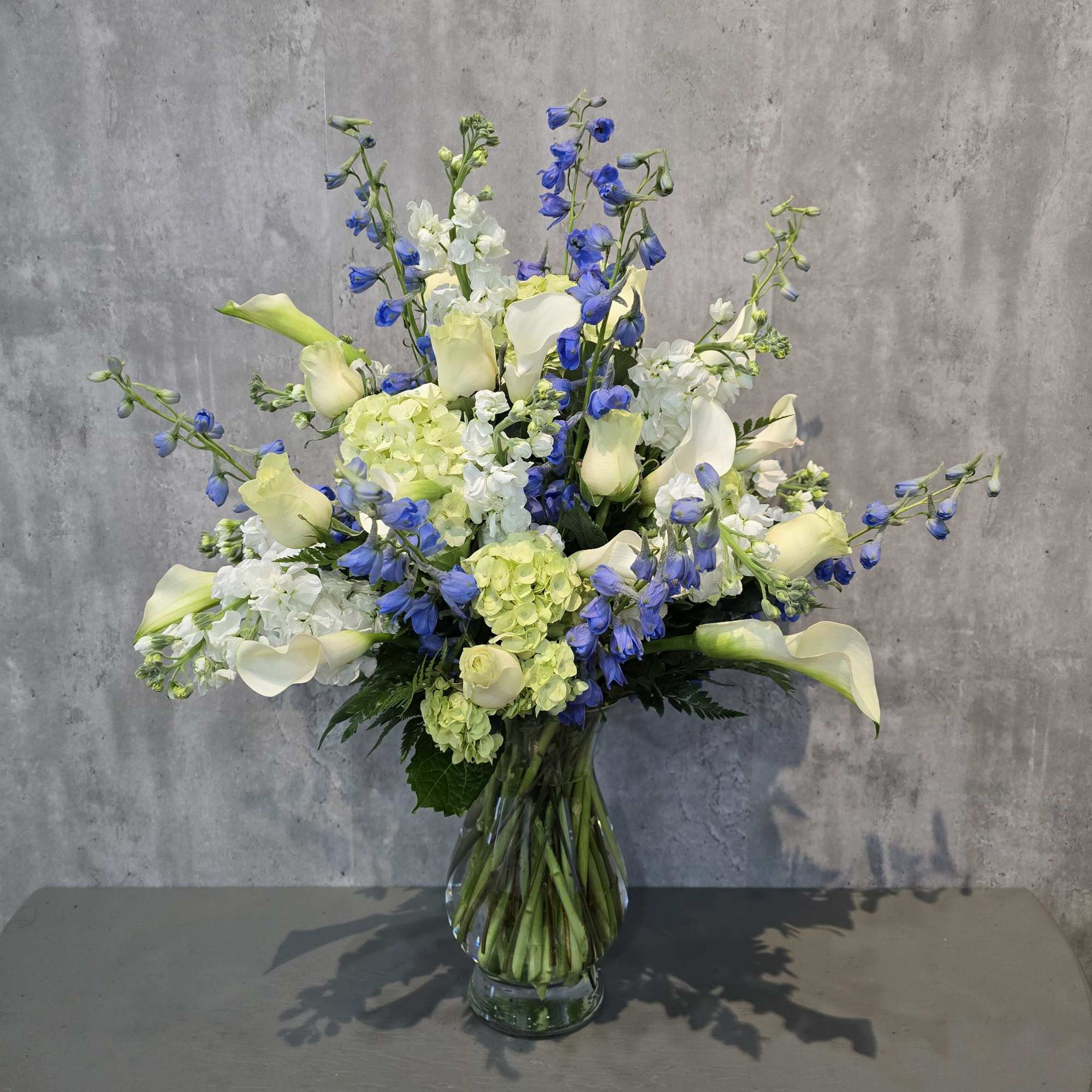 Tall bouquet of white calla lilies, blue flowers, and pale green hydrangeas in a glass vase