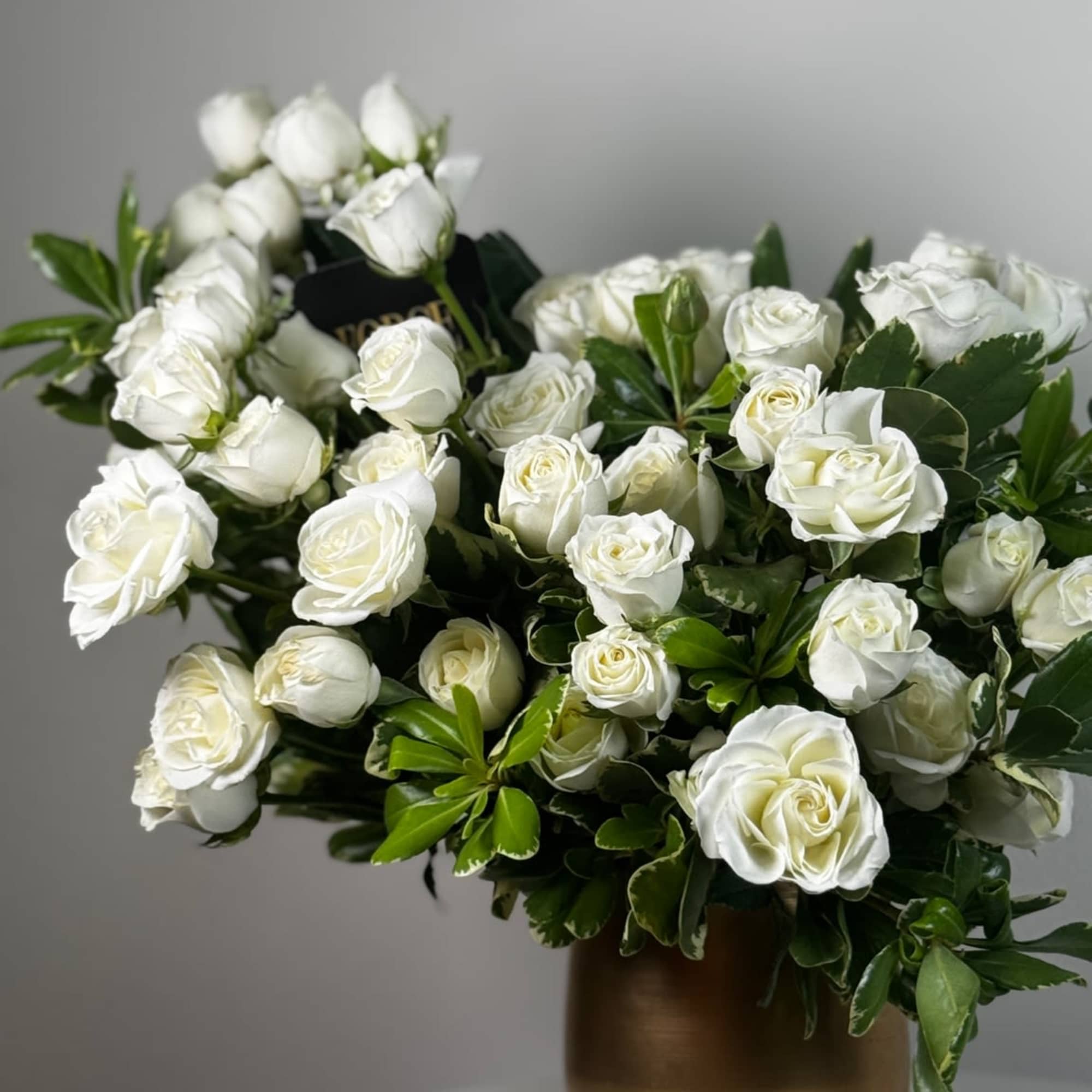 Bold white spray roses arranged with lush greenery in a matte-gold vase.