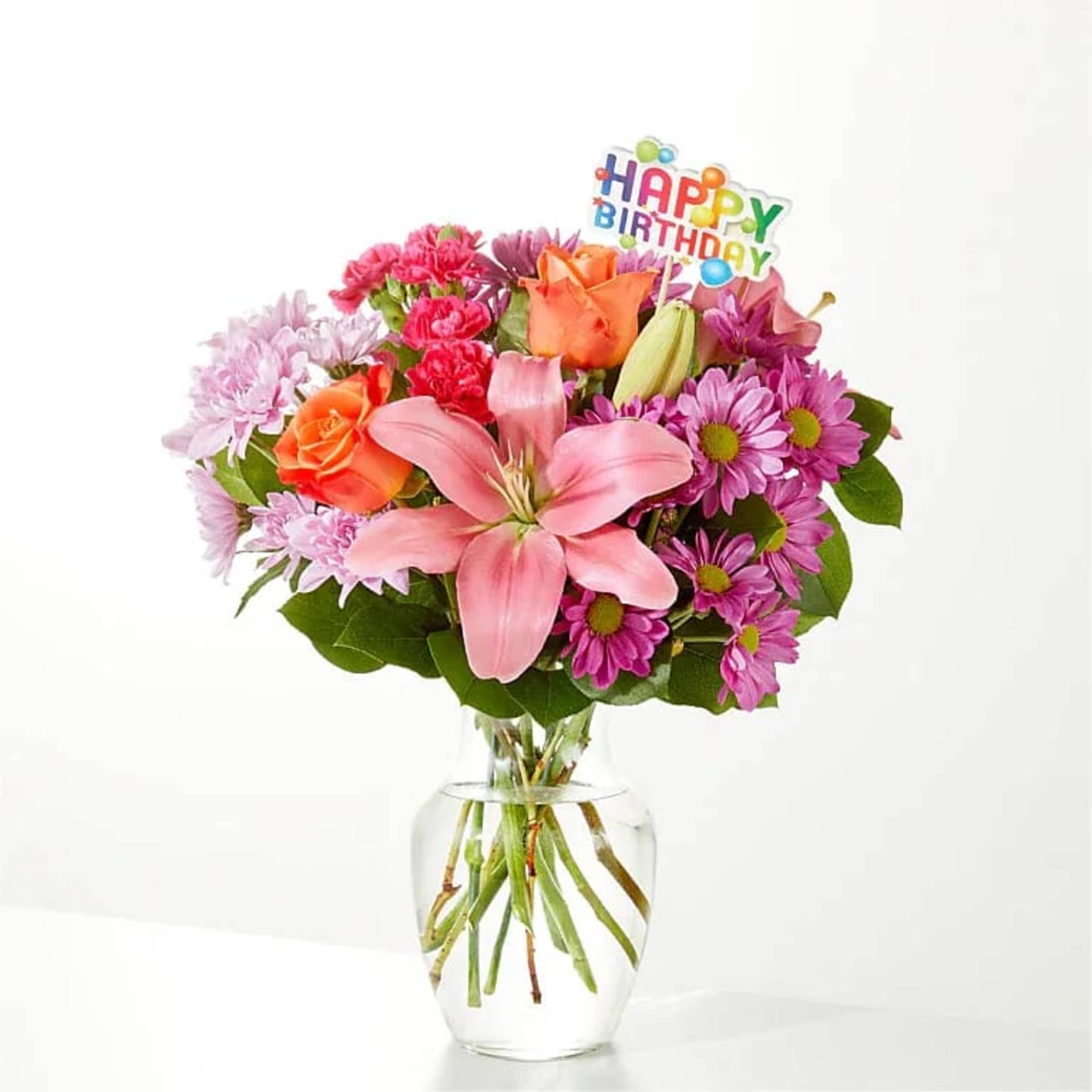 Bright mixed bouquet of pink lilies, orange roses, and purple mums in a clear vase with a Happy Birthday pick.