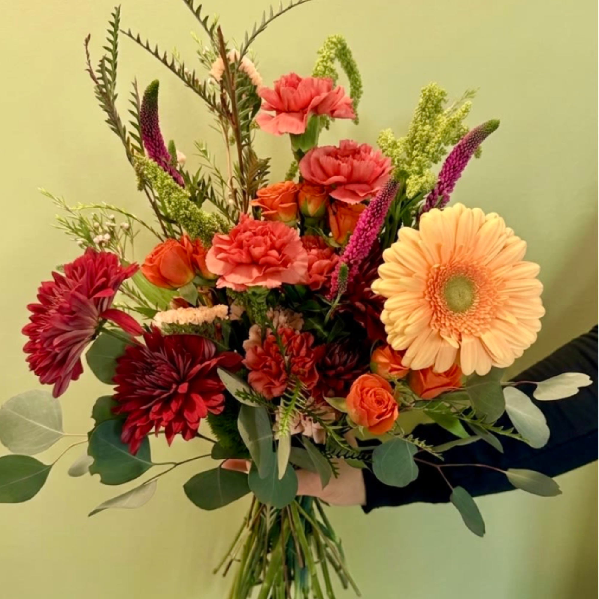 Hand-tied bouquet of orange gerbera, red mums, carnations, and roses with tall filler stems, held against a pale wall