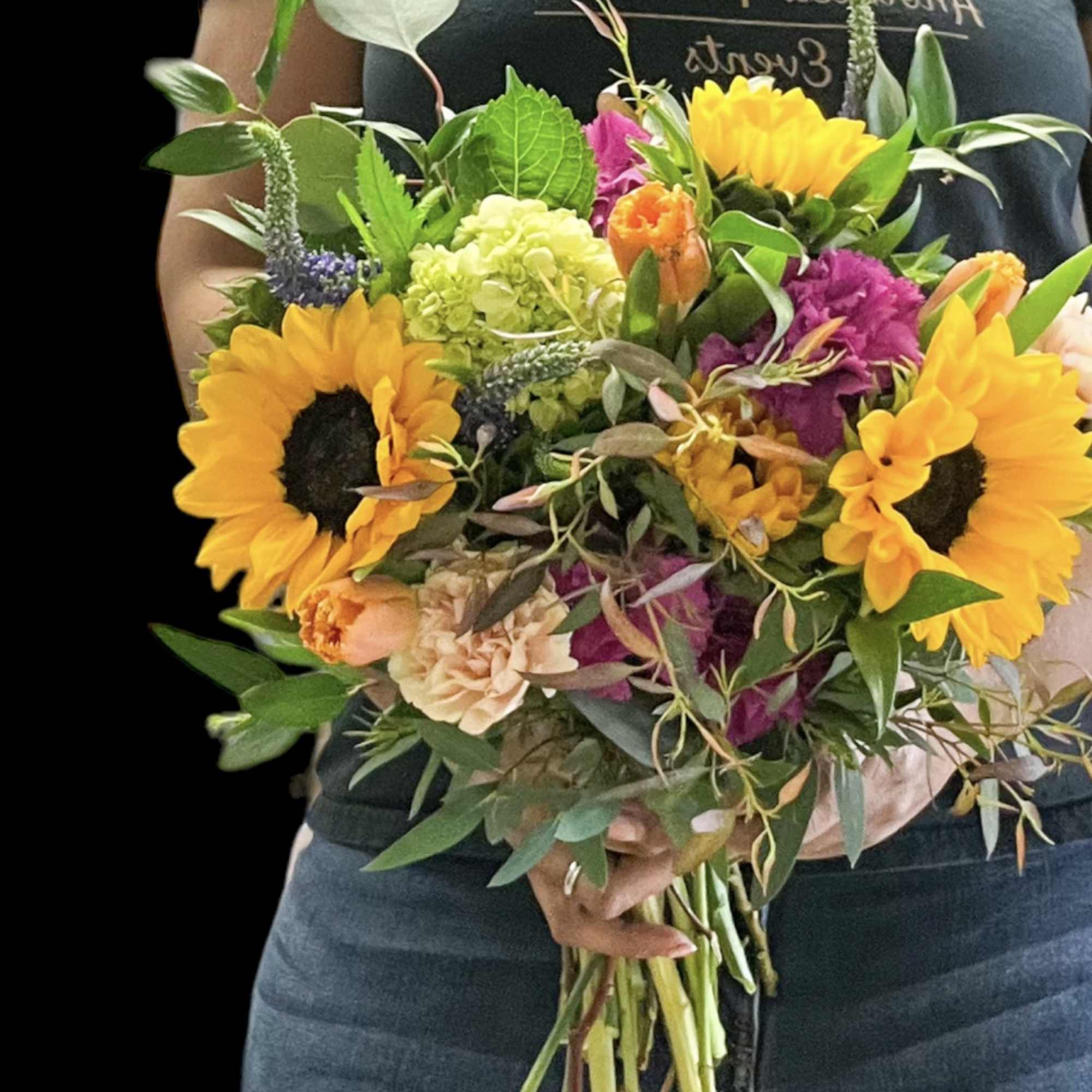 Hand-tied bouquet of yellow sunflowers and mixed bright flowers held by a person in a dark shirt