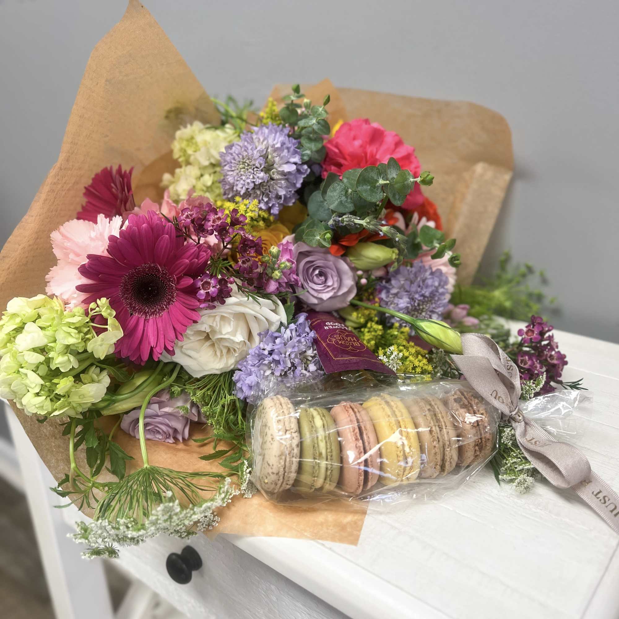 Colorful mixed bouquet with pink gerbera, roses, and pastel blooms wrapped in kraft paper with bag of macarons