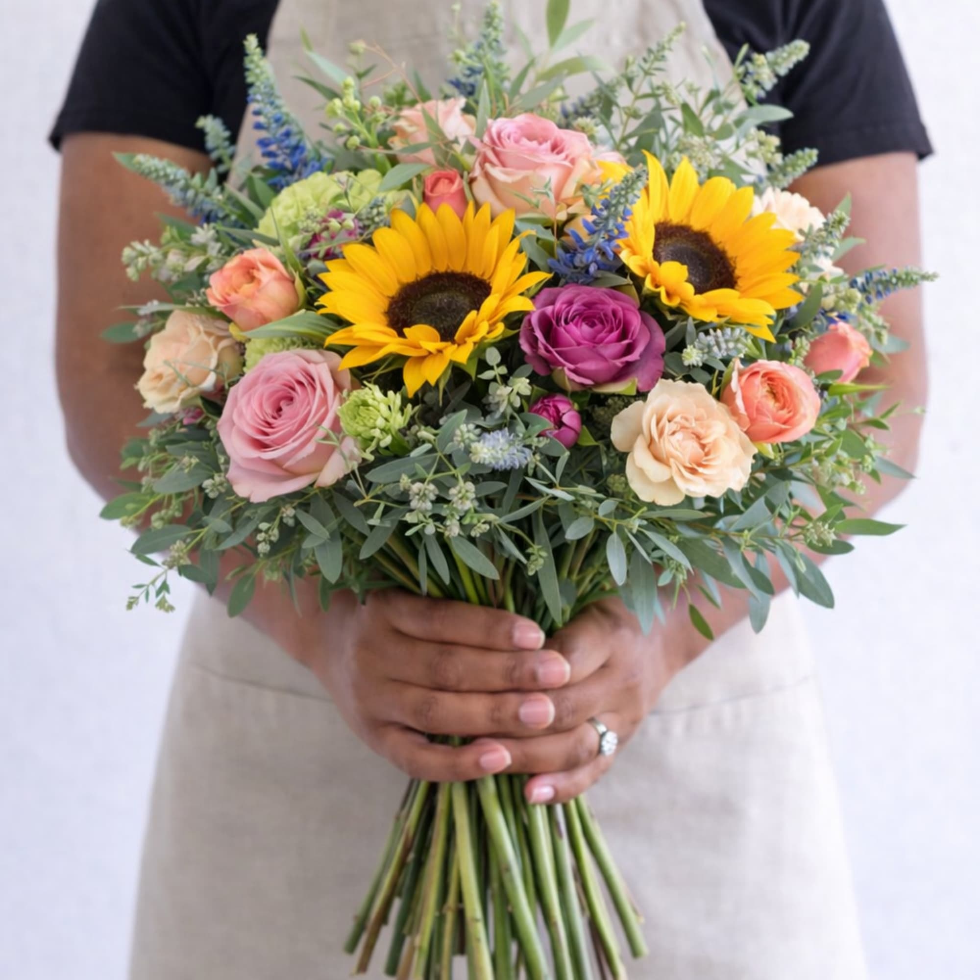 Hand-tied bouquet with sunflowers, pink and peach roses, and mixed blooms held by a person in an apron.