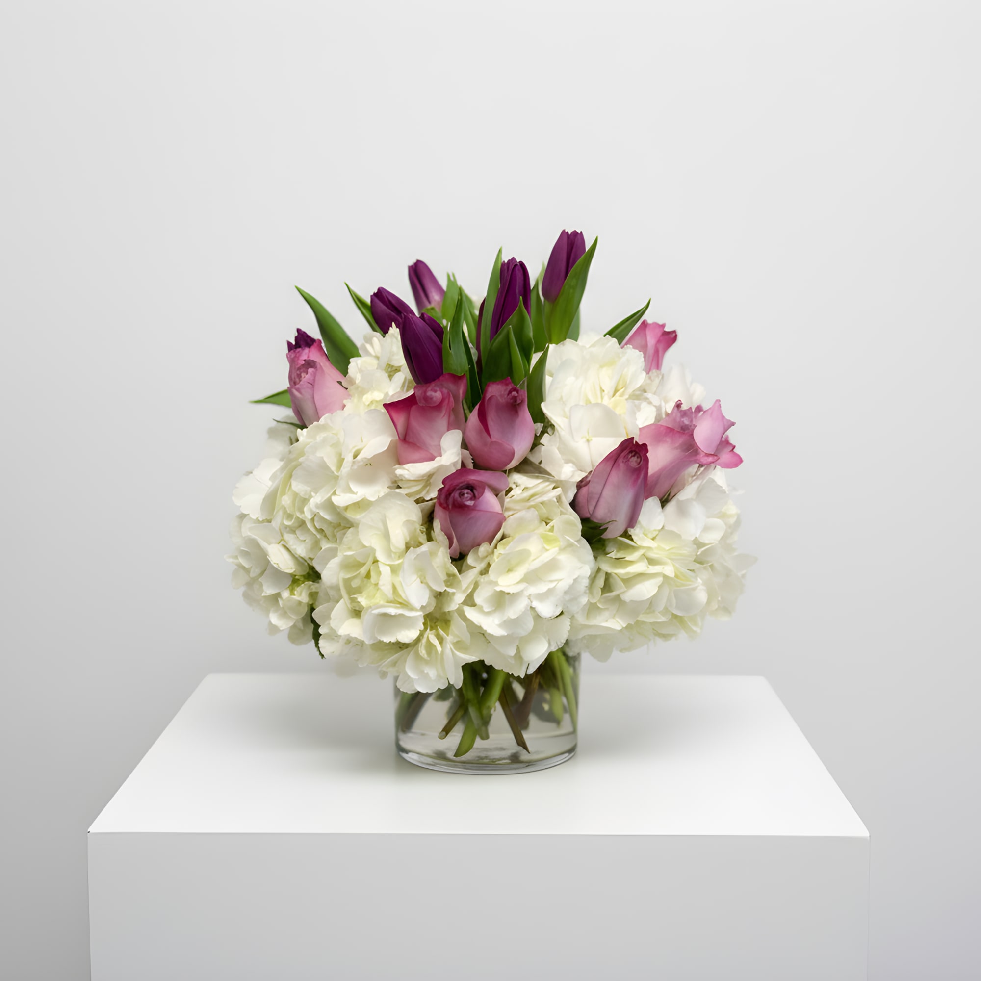 Low round arrangement of white hydrangeas with pink roses and purple tulips in a clear glass vase