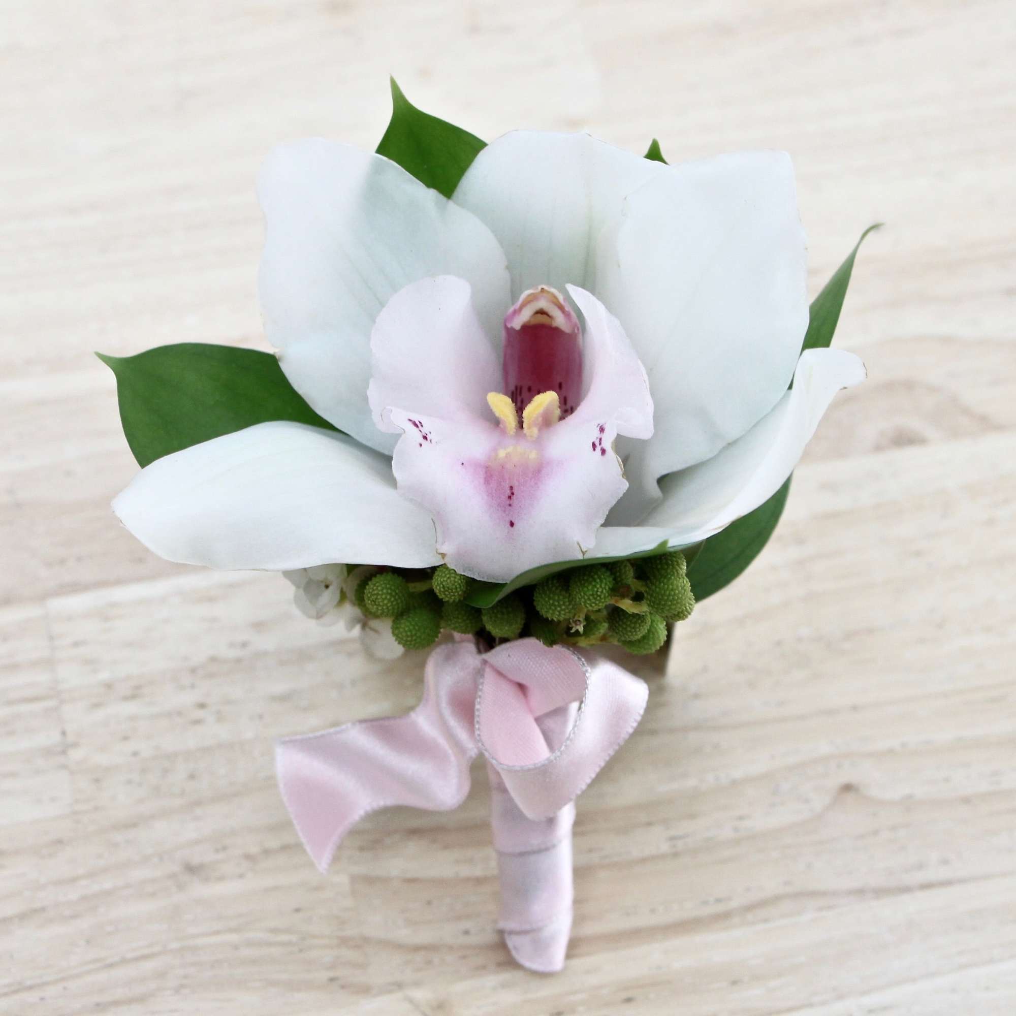 White and pink orchid boutonniere with green accents and pink ribbon on a light wood surface