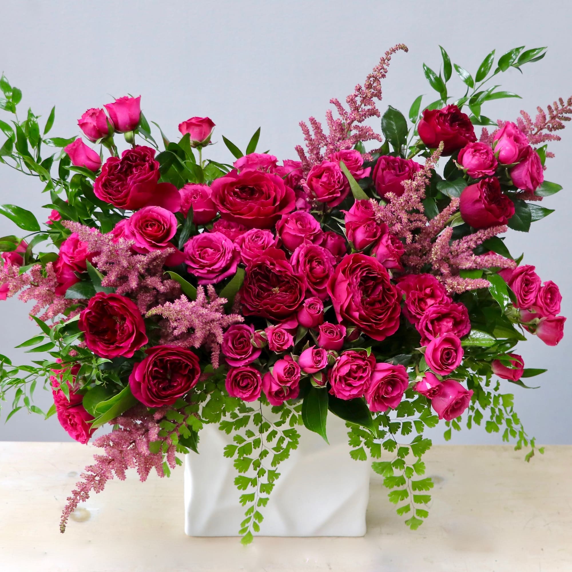 Wide hot pink rose arrangement with feathery pink accents and trailing ferns in a white rectangular vase