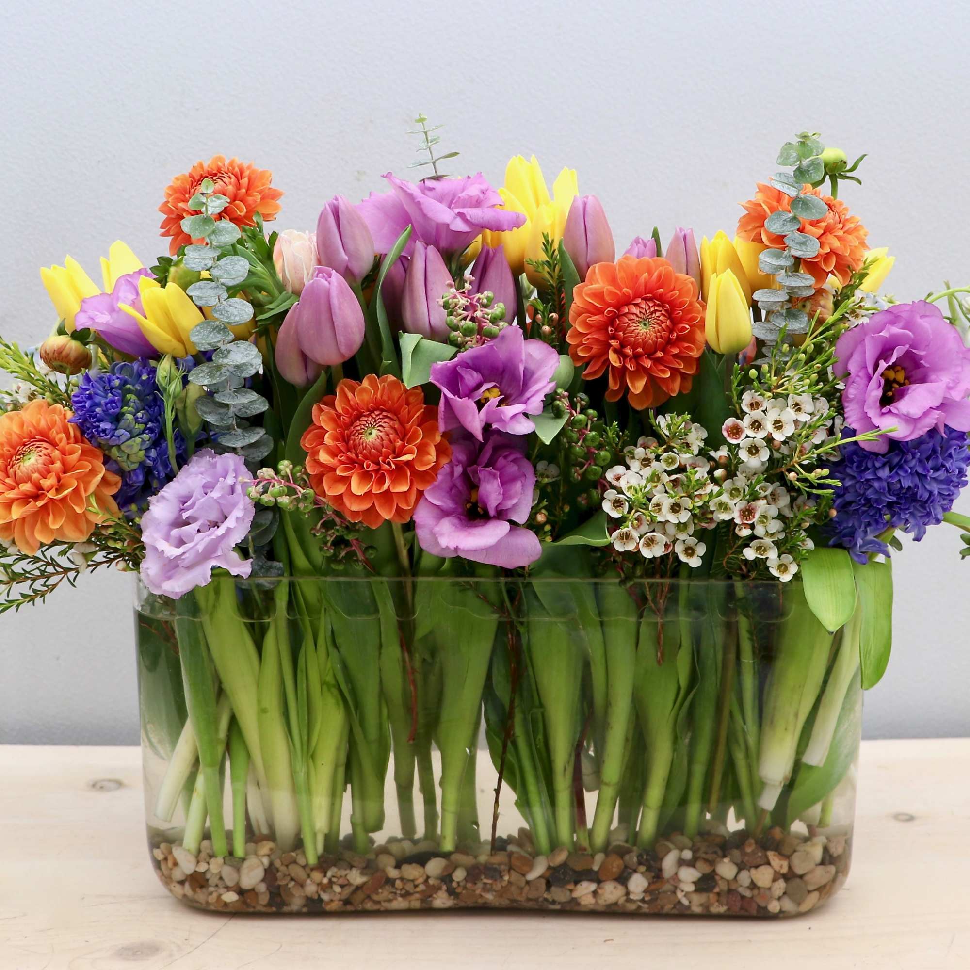 Low rectangular vase of orange dahlias, purple lisianthus, and yellow tulips with visible stems and pebbles.