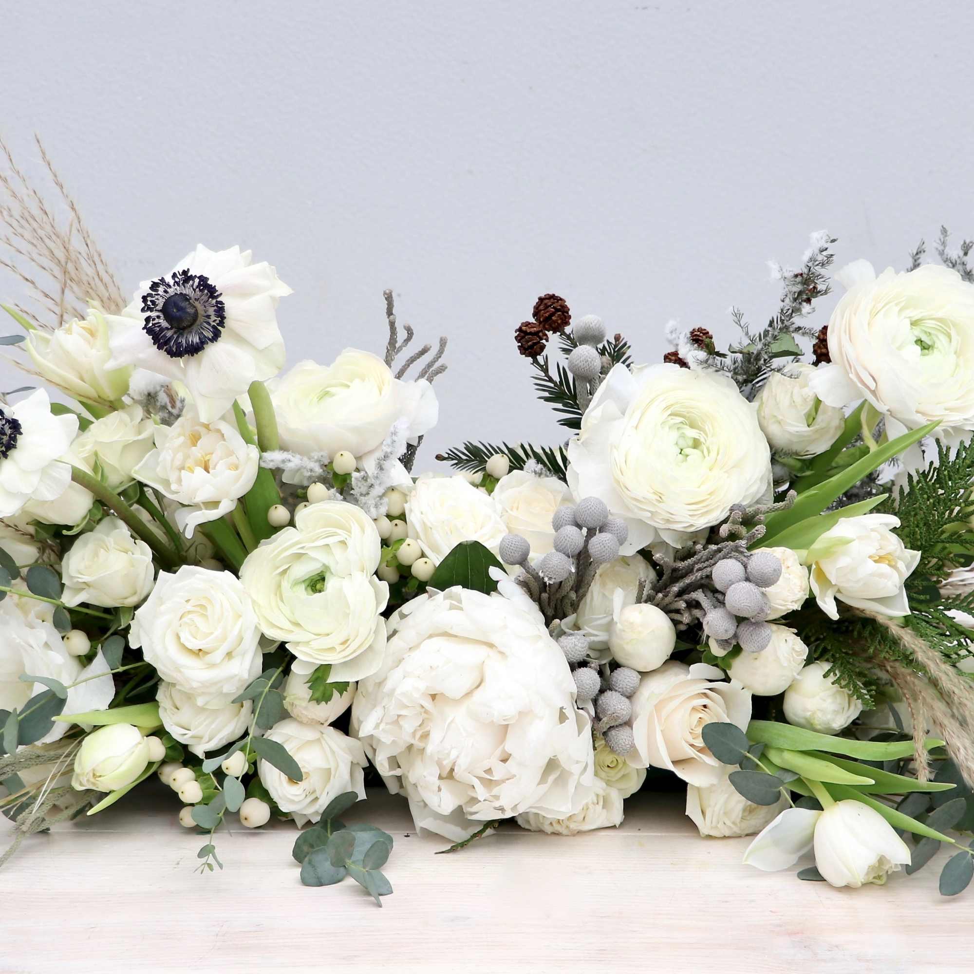 Low centerpiece of white ranunculus, roses, tulips and anemones with winter greenery and berries