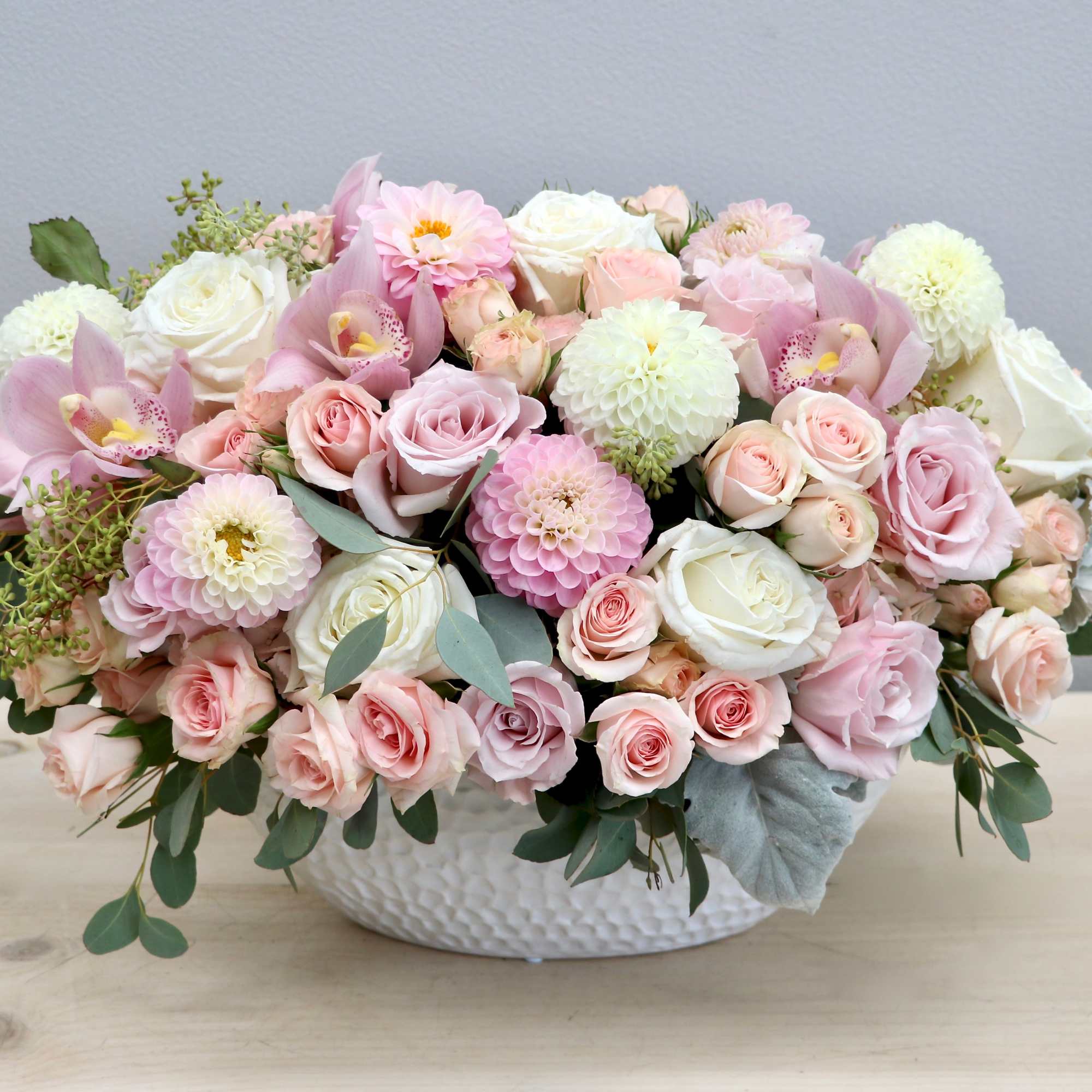 Low arrangement of pink and white roses, dahlias, and orchids in a white ceramic bowl