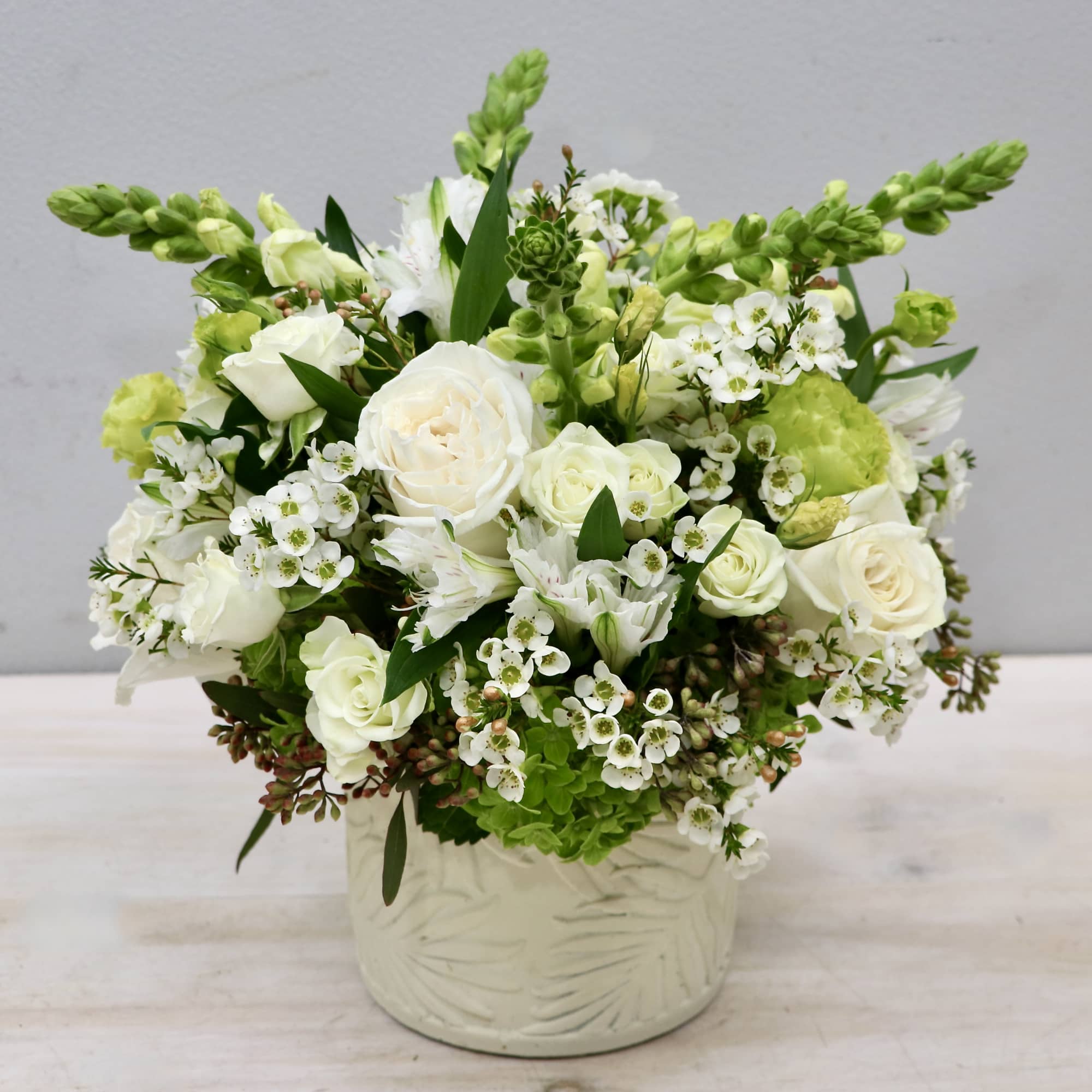 Low white and green floral arrangement with roses and mixed blooms in a textured white ceramic pot