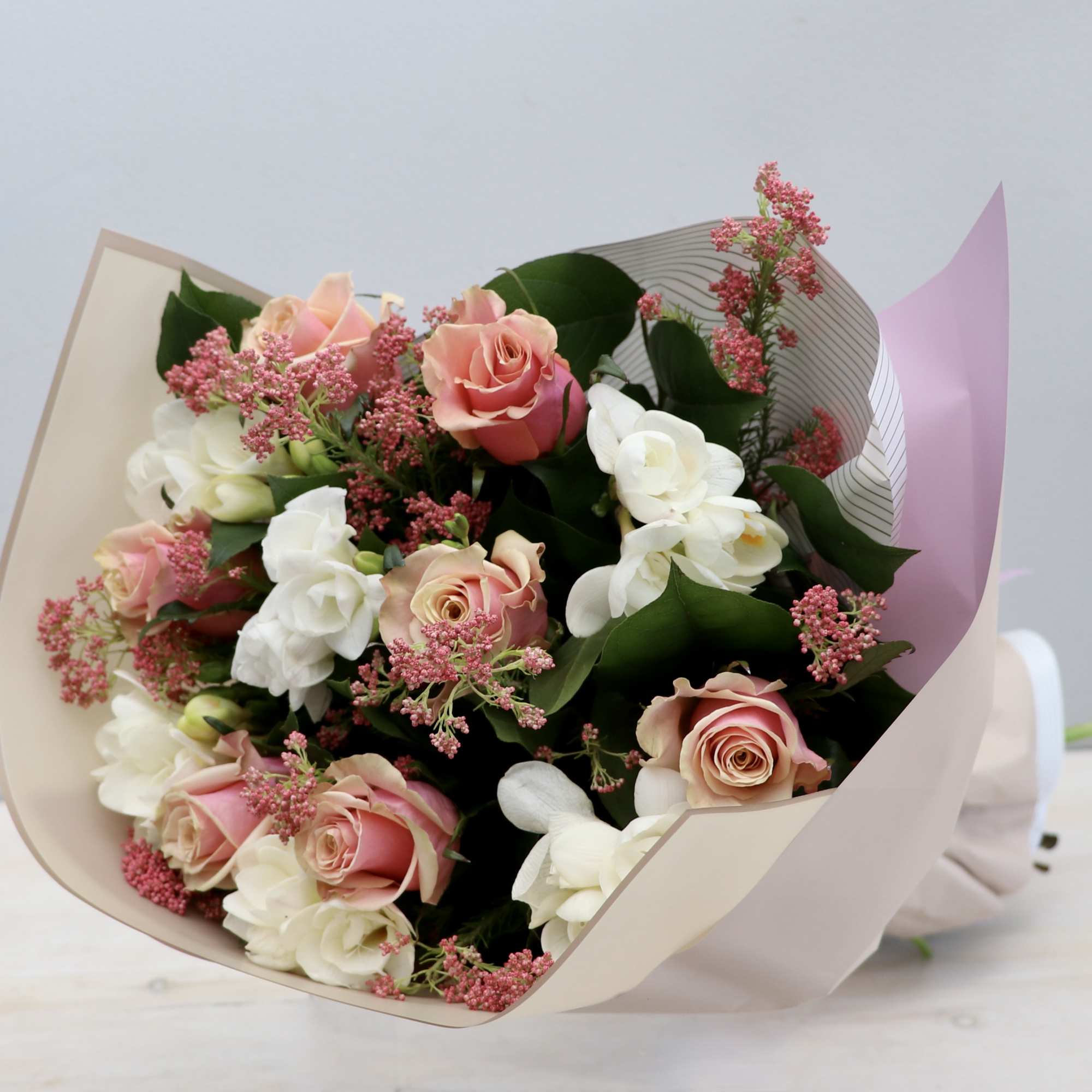 Hand-tied bouquet of pink roses, white flowers, and pink filler blooms wrapped in pastel paper