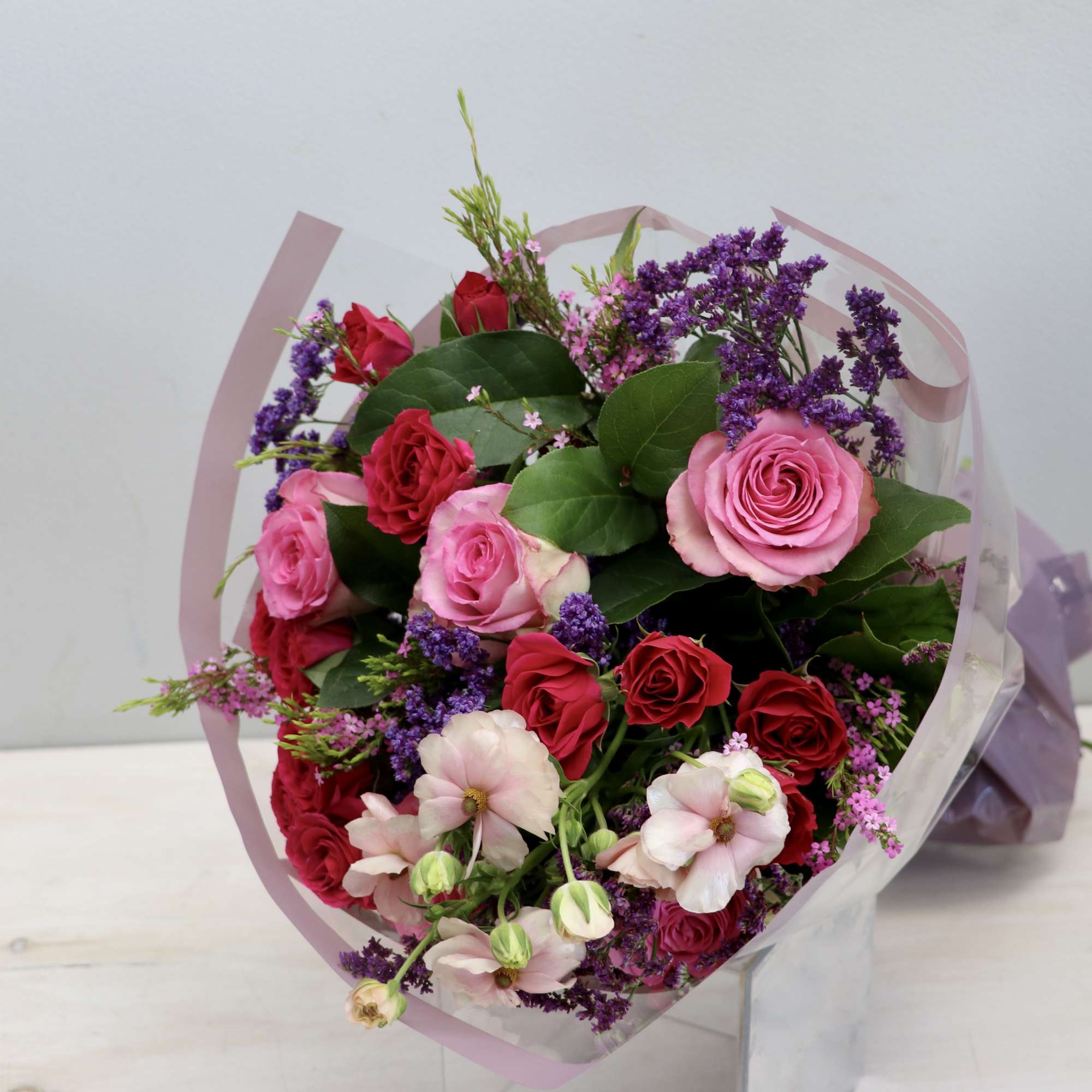 Hand-tied bouquet of pink and red roses with purple filler flowers wrapped in pink cellophane