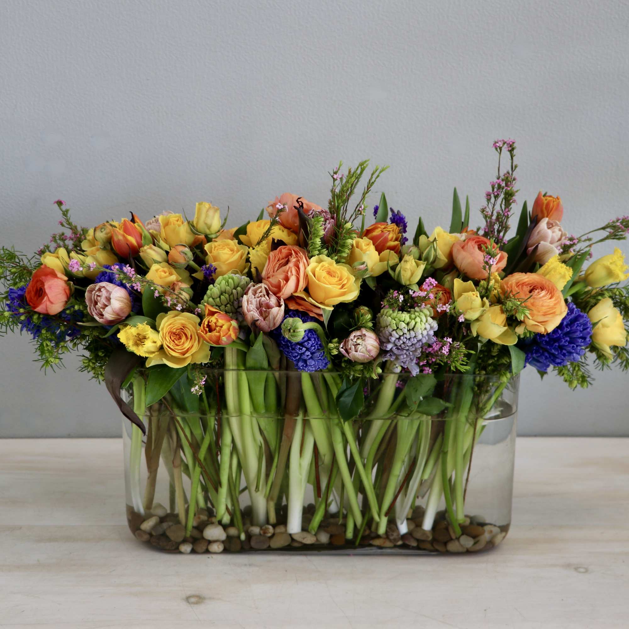 Low rectangular arrangement of yellow, orange, and purple flowers in a clear glass vase
