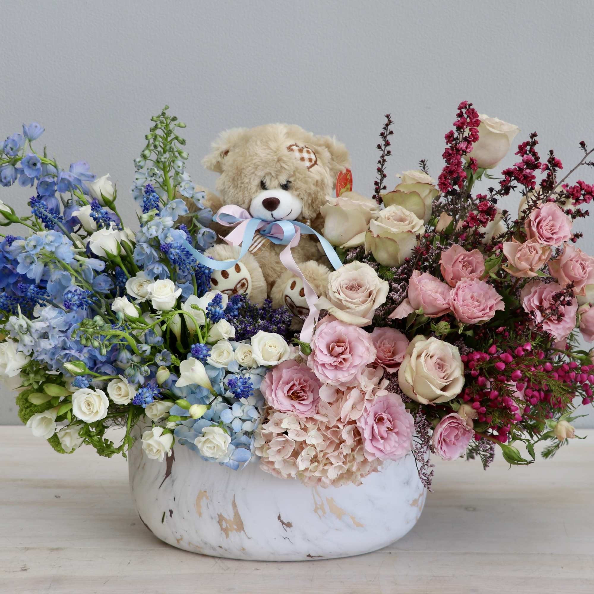 Large pink and blue flower arrangement in a white bowl with a plush teddy bear in the center