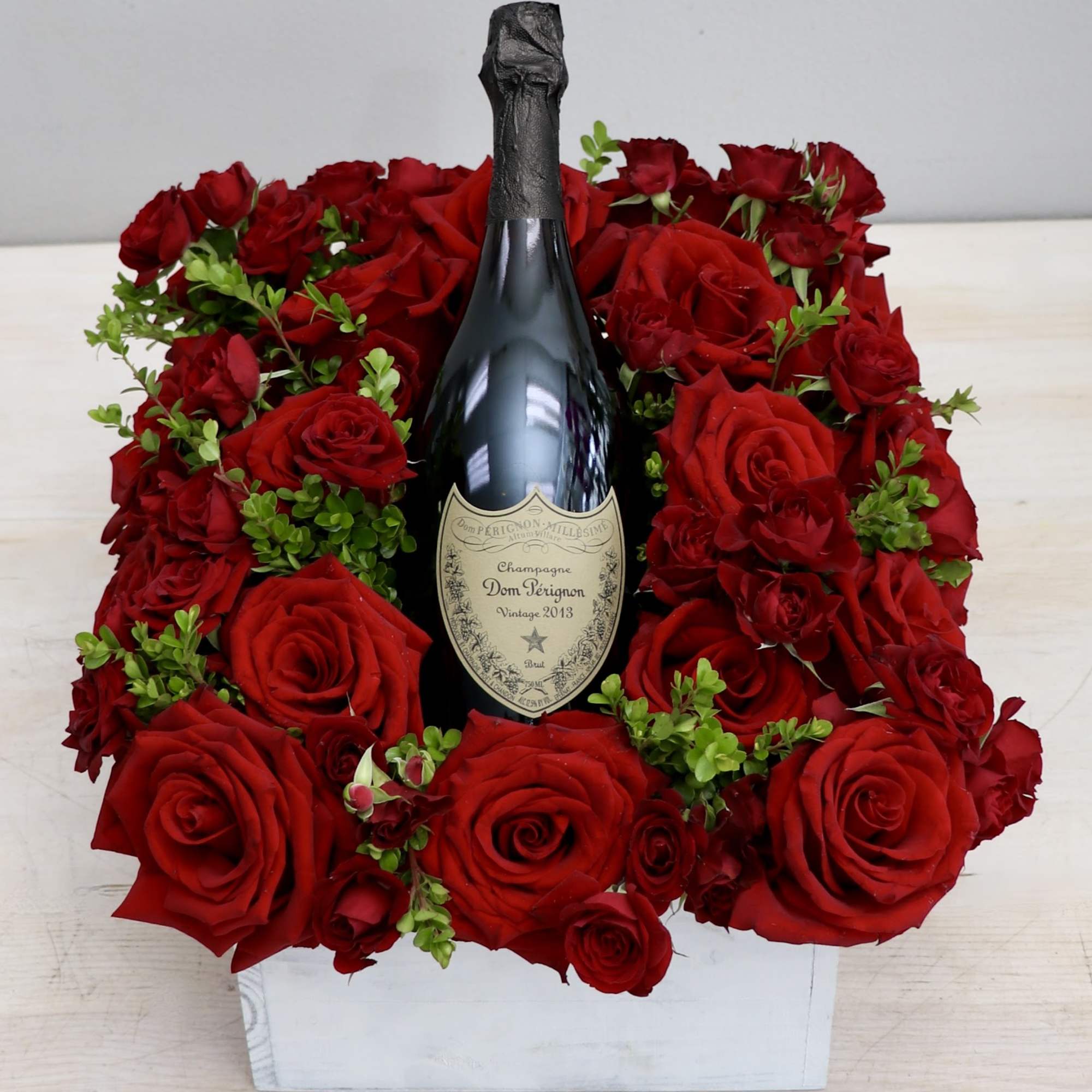 Square arrangement of red roses surrounding a champagne bottle in a white wooden box