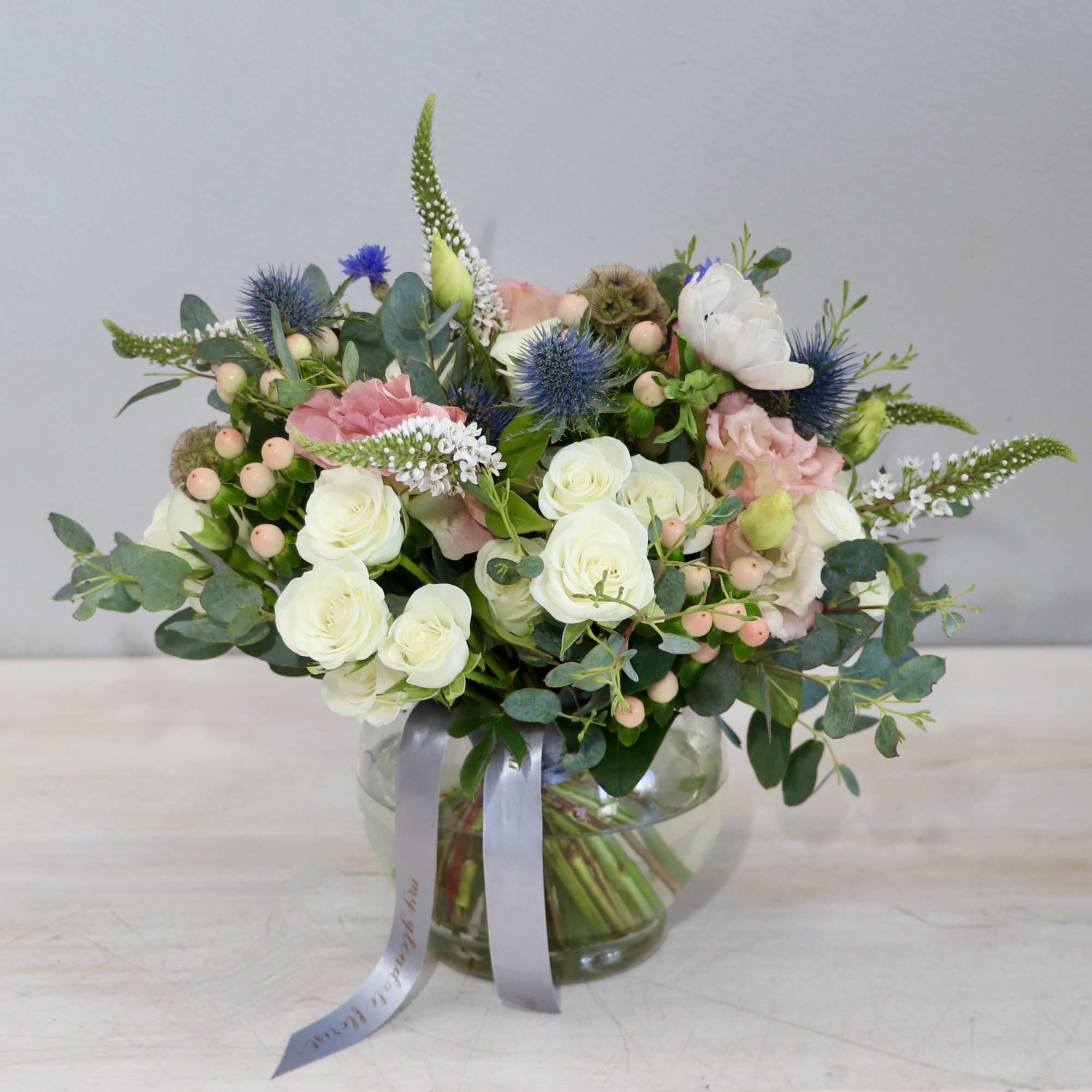 Low pastel arrangement of white roses, blush flowers, and blue thistle in a clear round vase with a gray ribbon.